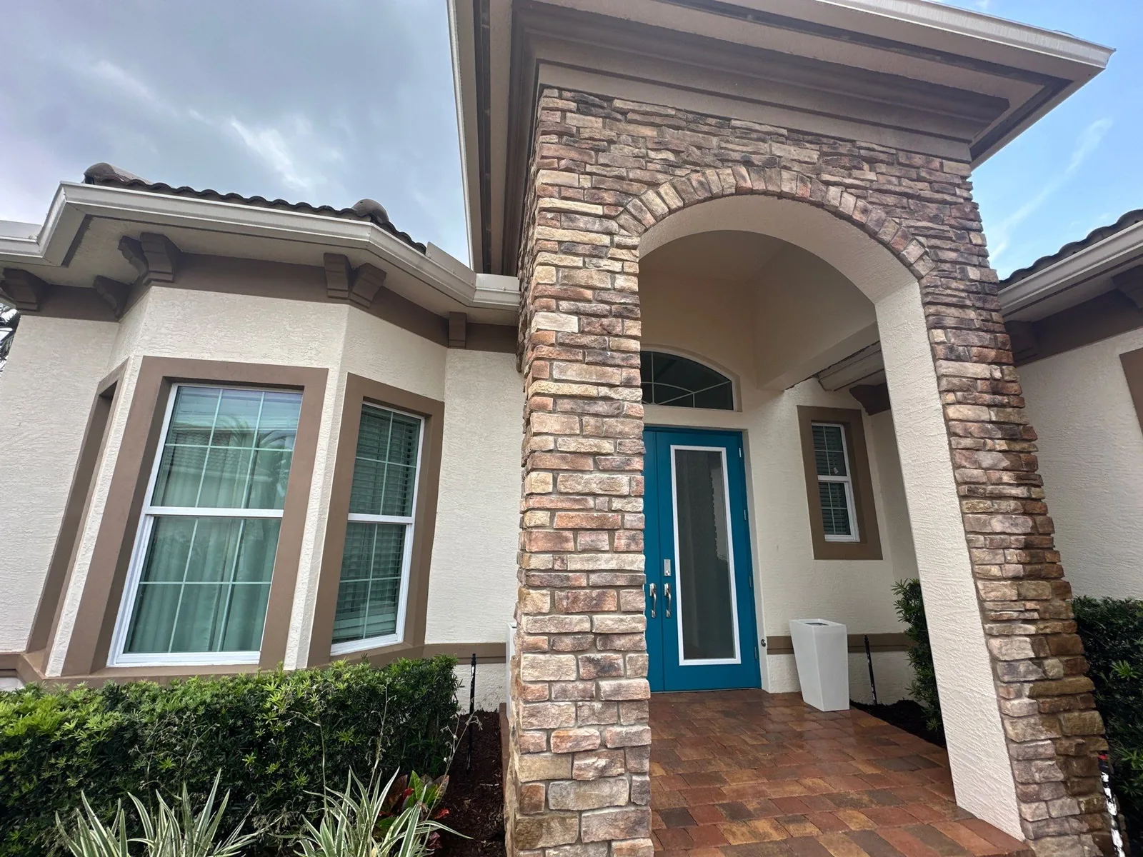 Exterior Painting in Port Saint Lucie, Florida. Front entry of a home with tan stucco, brown window and trim paint, a stone-veneer arched portico and a teal front door.