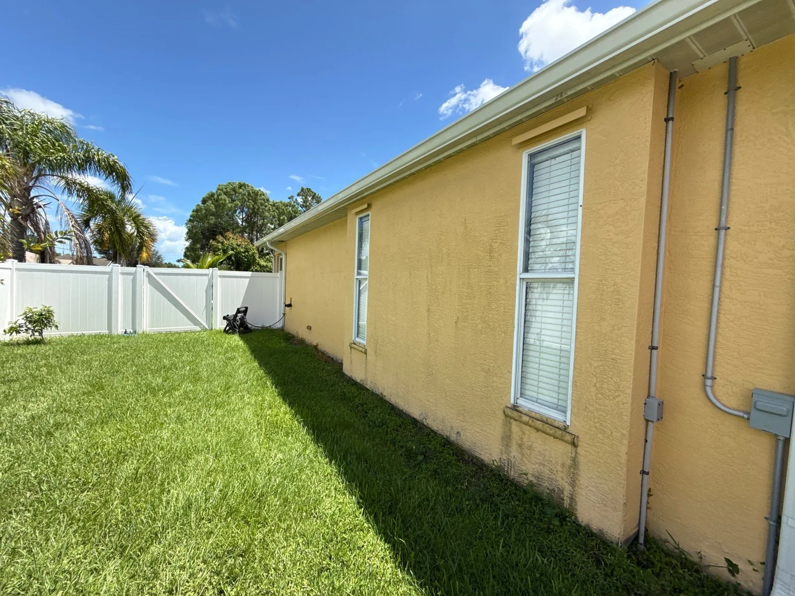 Exterior Painting in Port Saint Lucie, Florida. Yellow stucco house side elevation with freshly painted white window trim, green lawn and white vinyl fence under blue sky.