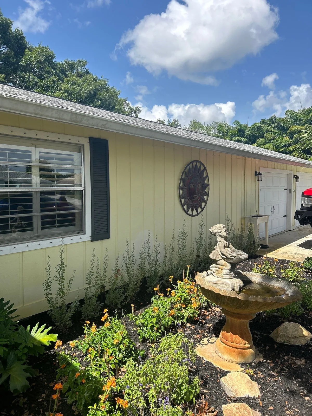 Exterior Painting in Port Saint Lucie, Florida. Single-storey home with pale yellow painted siding, black window shutters and white trim, with a garden and stone birdbath in front.