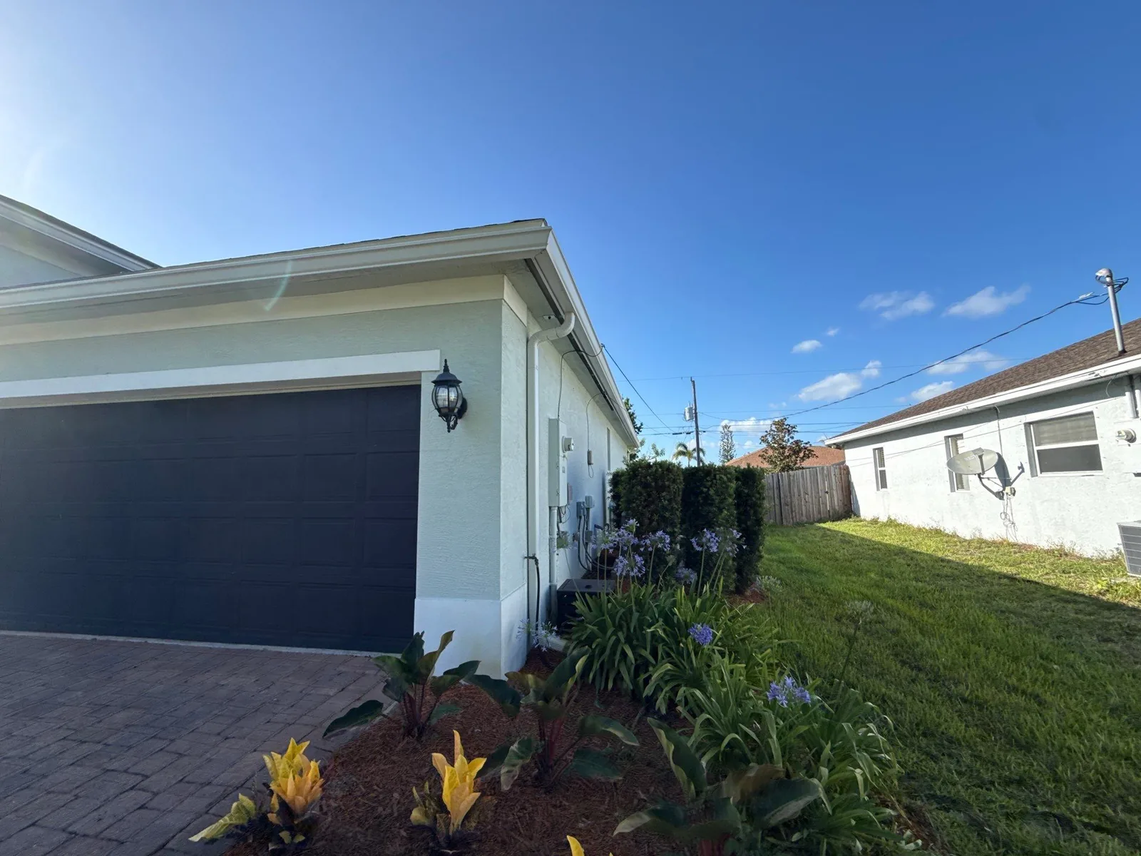 Exterior Painting in Port Saint Lucie, Florida. Garage end of a single-storey white stucco house with a dark garage door, coach light and white fascia, a paver driveway and a flowering planting bed, under a clear blue sky; overhead utility lines and a neighbouring house are visible at right.