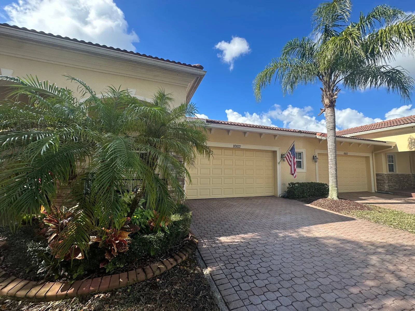 Exterior Painting in Port Saint Lucie, Florida. Single-storey home painted pale yellow with a matching garage door, paver driveway and palm plantings under a blue sky.