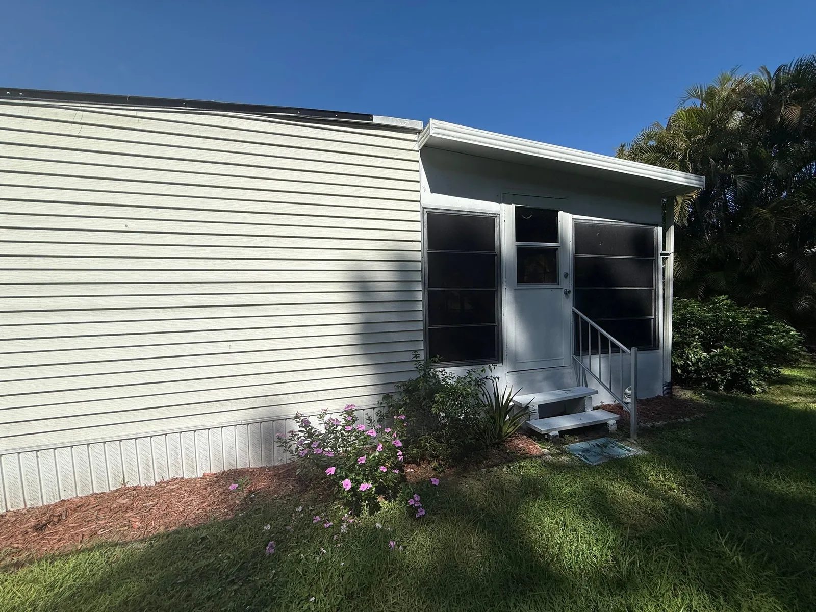 Exterior Painting in Port Saint Lucie, Florida. Corner of a single-storey home with white horizontal siding and trim, screened porch with entry steps and railing, flowering shrub and lawn under a clear sky.