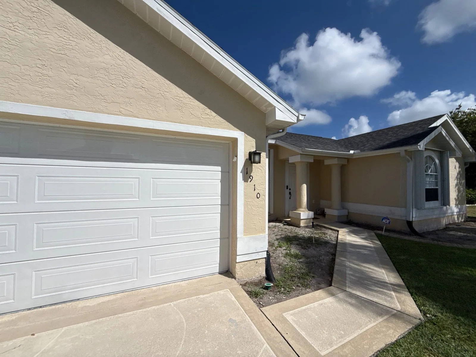 Exterior Painting in Port Saint Lucie, Florida. Tan stucco single-storey home with a white sectional garage door, white trim and a columned front entry, shot from the driveway.