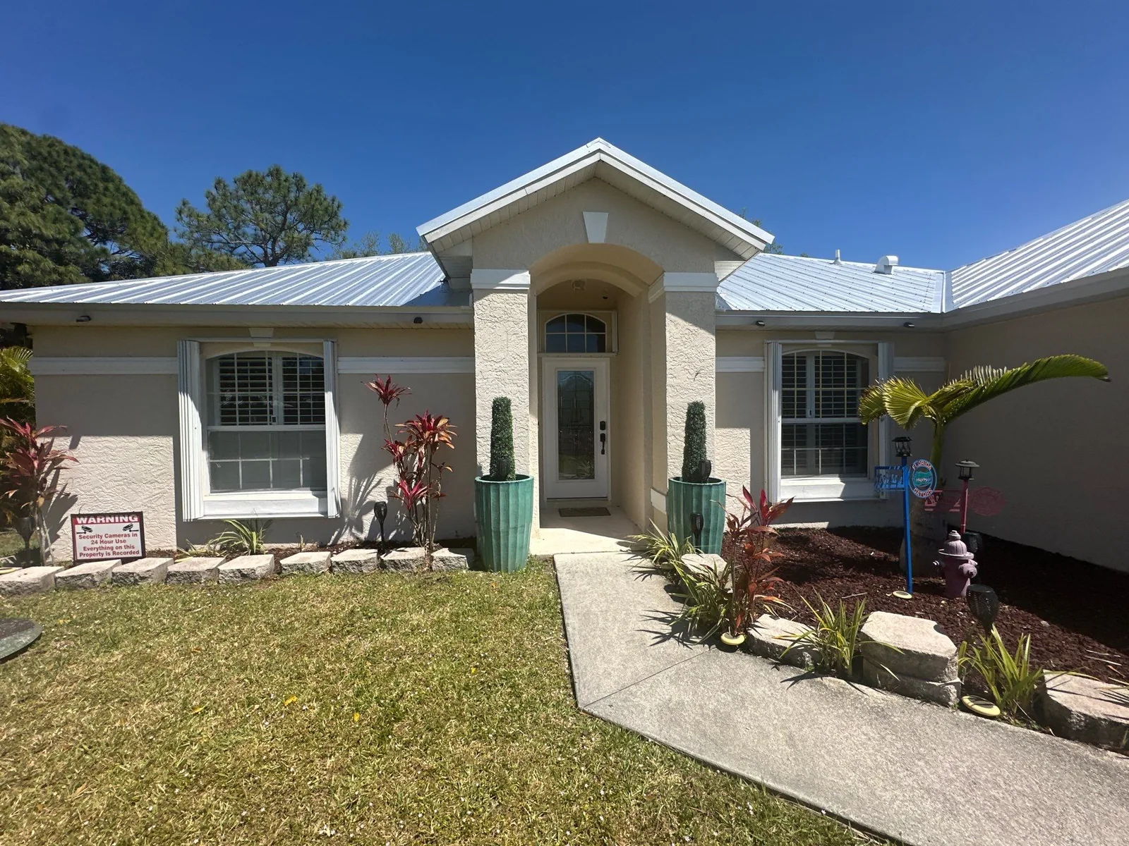 Exterior Painting in Port Saint Lucie, Florida. Single-storey stucco home front elevation, cream walls with white trim and arched entry, metal roof and landscaped beds.