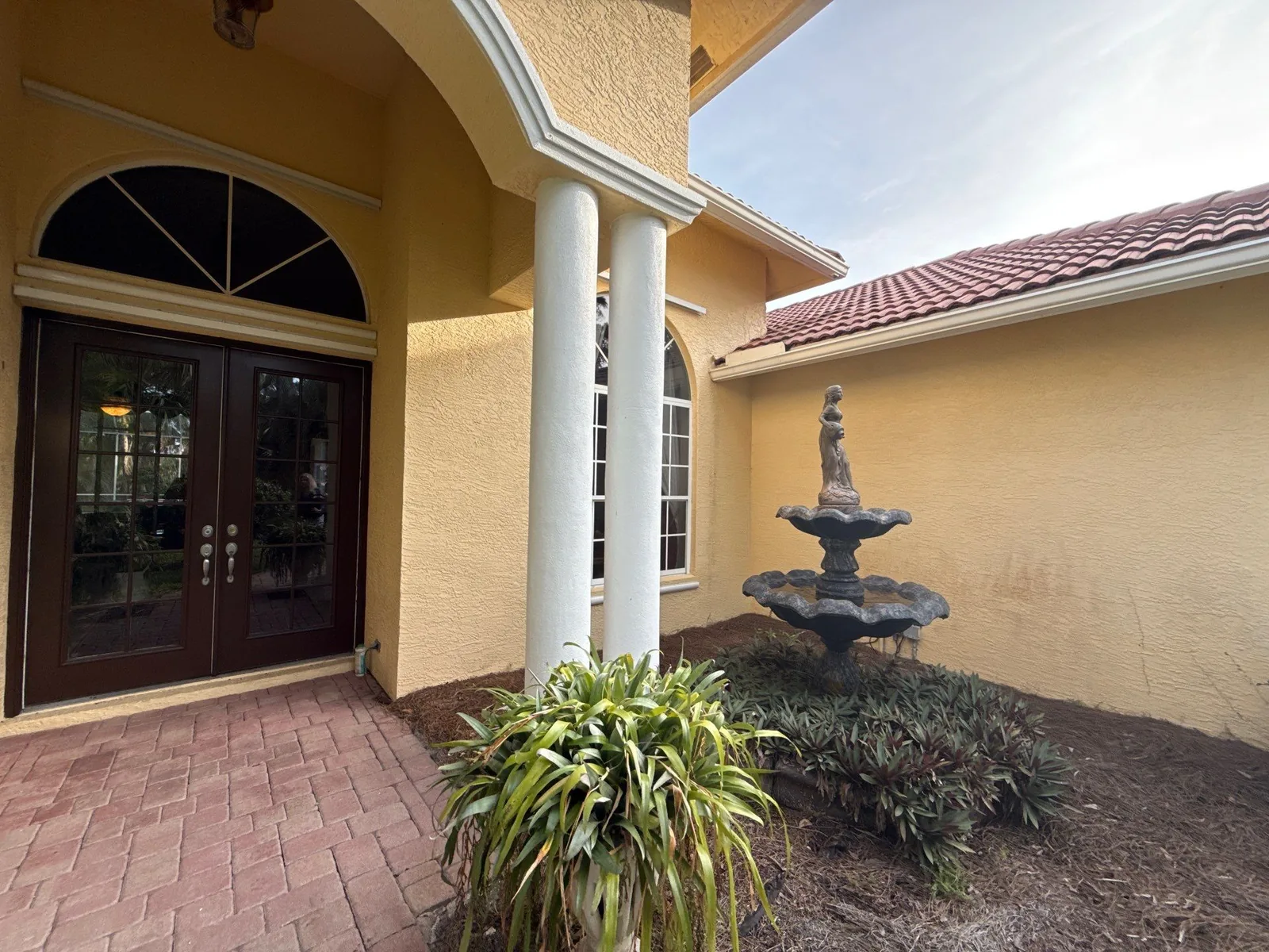 Exterior Painting in Port Saint Lucie, Florida. Yellow stucco home entry with a white column and trim, dark wood double doors, arched window, barrel tile roof and a tiered stone fountain.