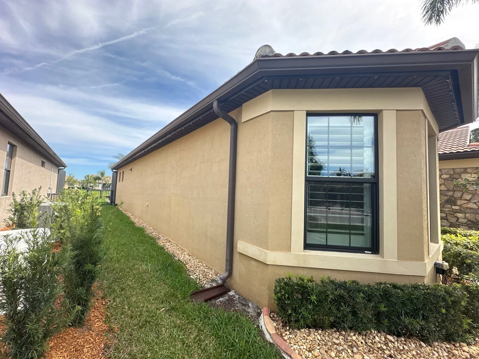 Exterior Painting in Port Saint Lucie, Florida. Side elevation of a single-storey tan stucco home with cream window surround, tile roof and trimmed hedges.