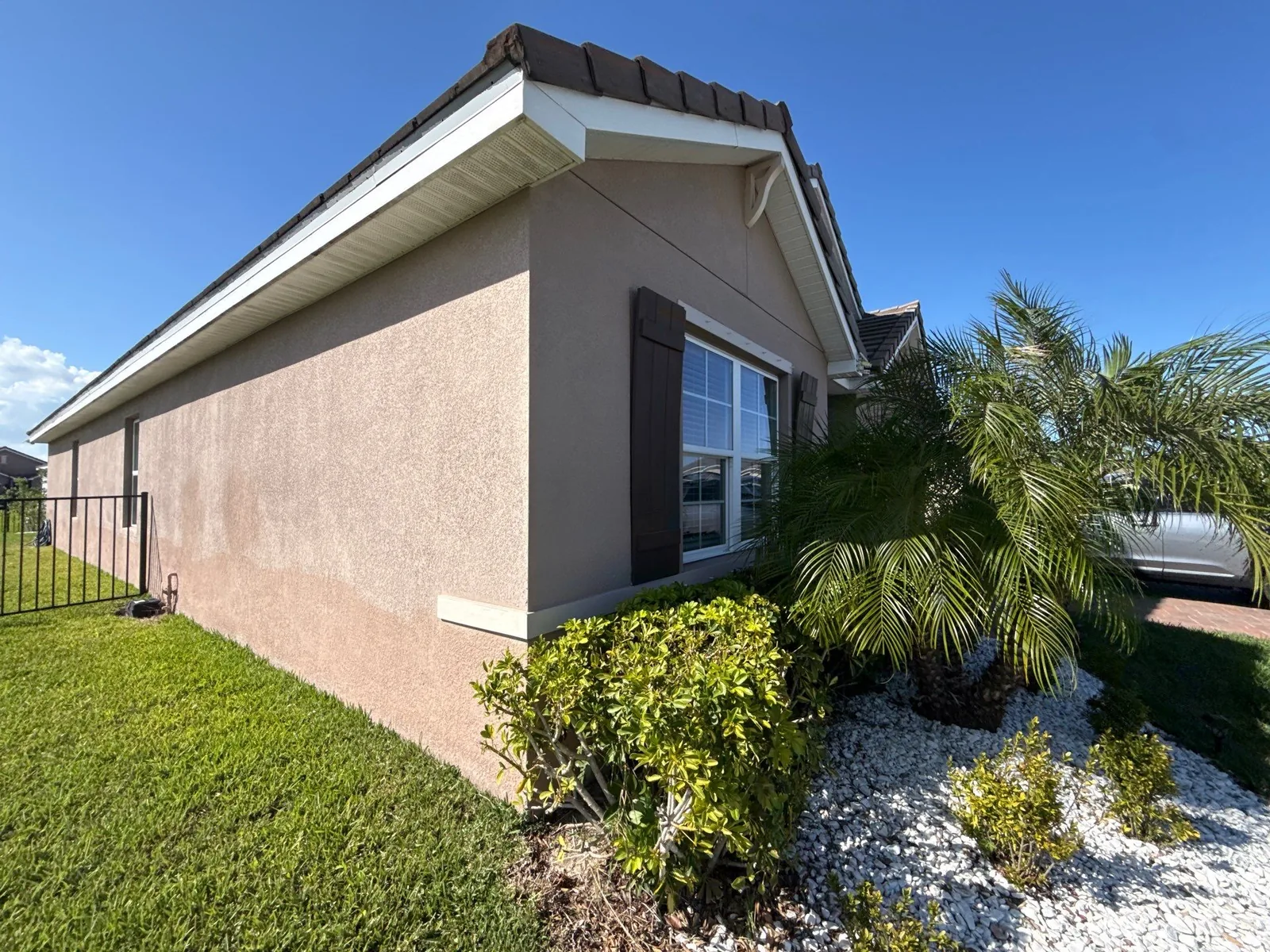 Exterior Painting in Port Saint Lucie, Florida. Corner of a single-storey stucco house painted warm taupe with white fascia and soffit, dark brown window shutters, shrubs and a palm in a white rock bed.