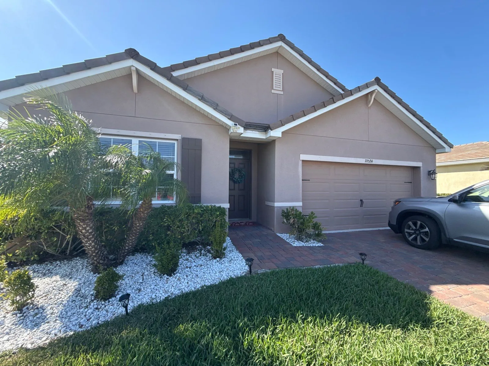 Exterior Painting in Port Saint Lucie, Florida. Front elevation of a single-storey stucco home in taupe with white trim, a brown garage door, paver driveway and landscaped rock beds.