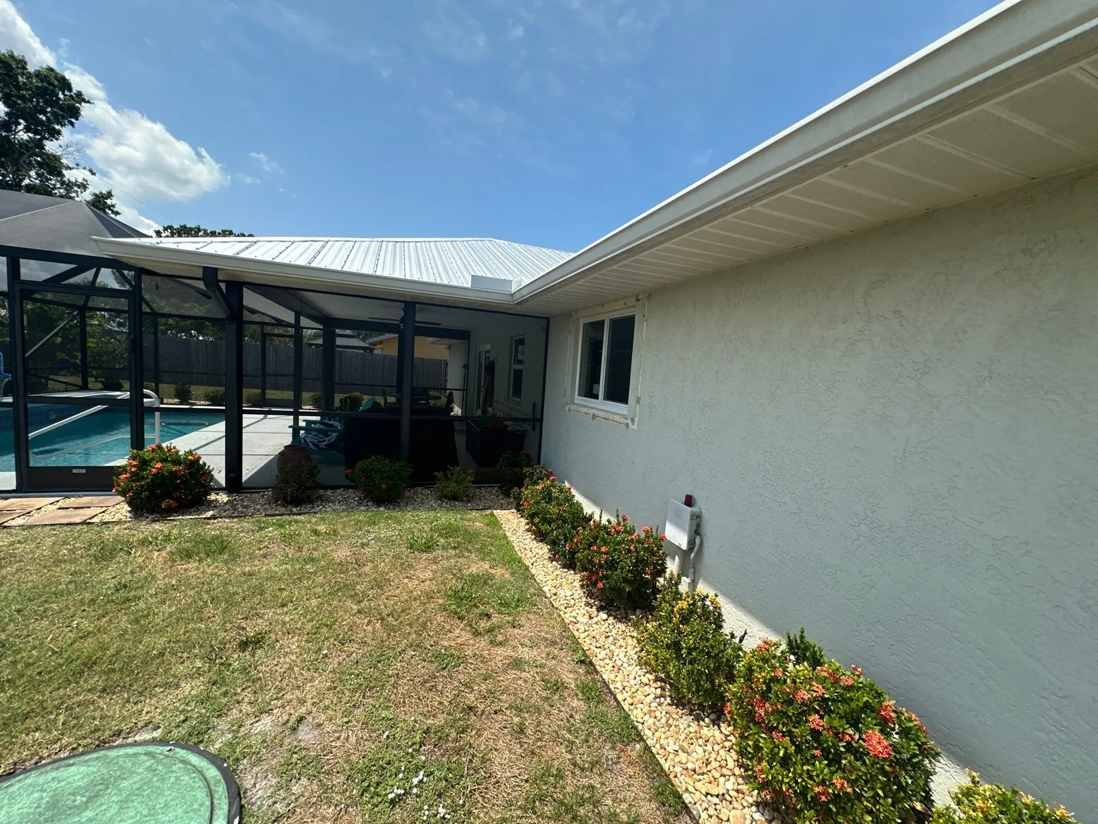 Exterior Painting in Port Saint Lucie, Florida. Side elevation of a single-storey house with white stucco walls and white soffit, metal roof and adjoining pool screen enclosure.