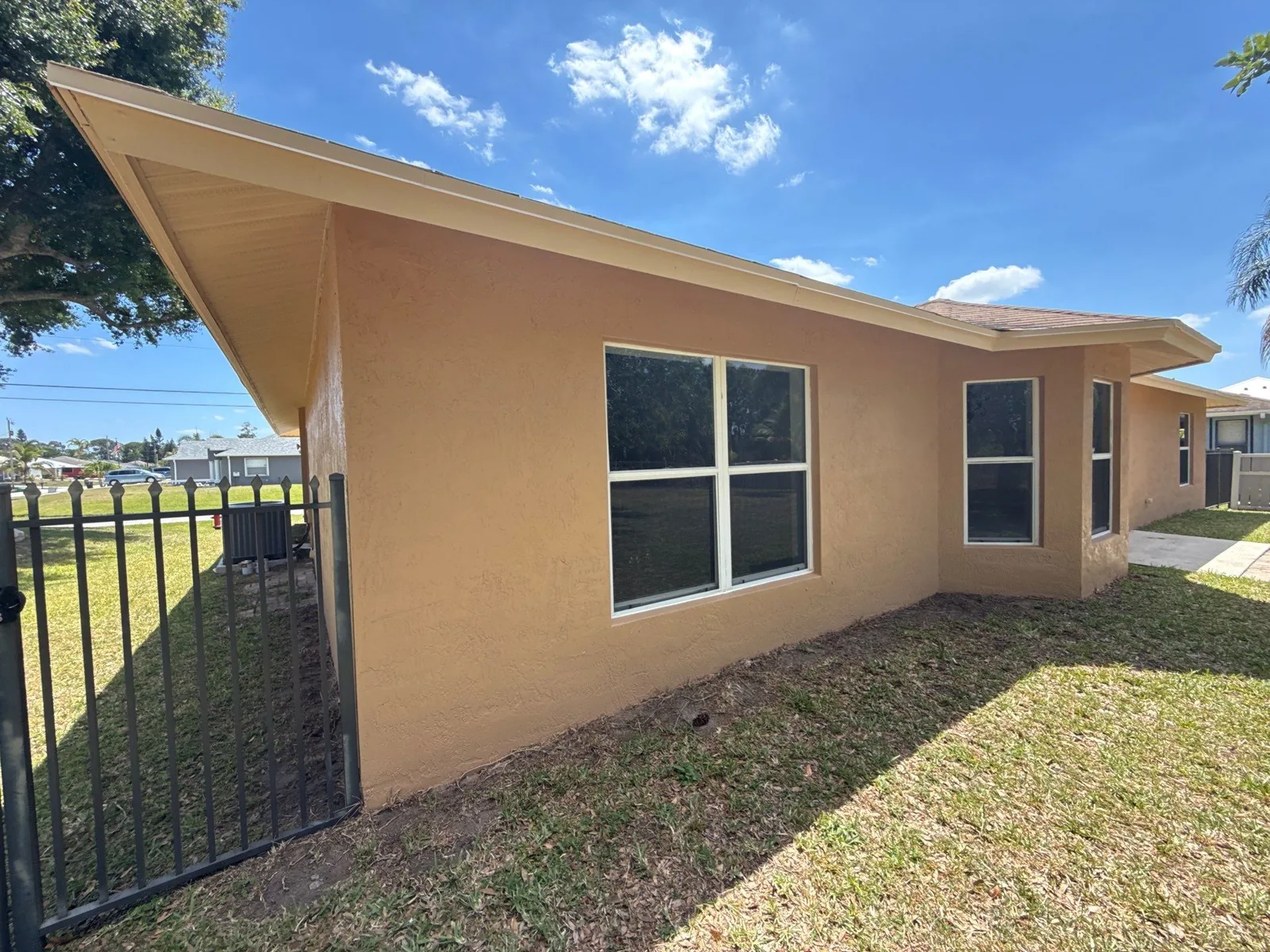 Exterior Painting in Port Saint Lucie, Florida. Corner view of a single-storey tan stucco house with white-trimmed windows and white soffit, green lawn and blue sky.