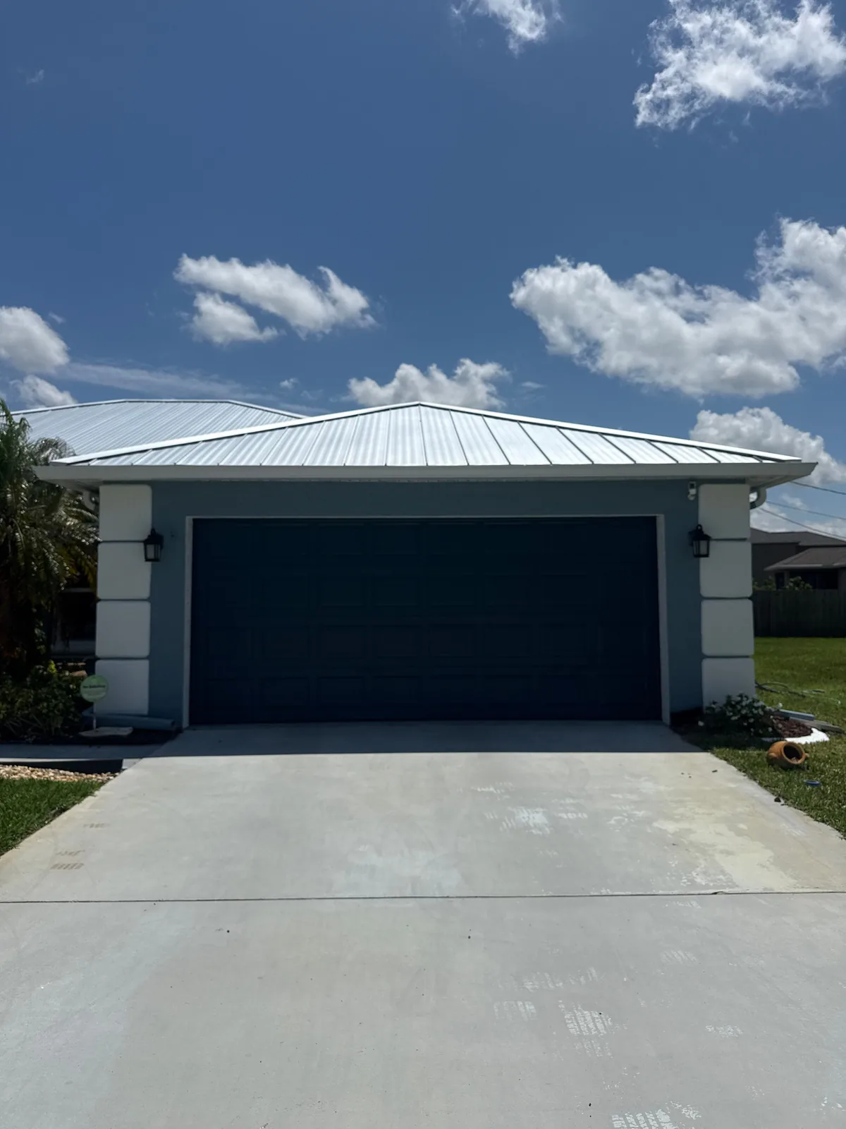 Exterior Painting in Port Saint Lucie, Florida. Two-car garage front with a dark teal garage door, pale blue-grey stucco walls and a white metal roof above a concrete driveway.