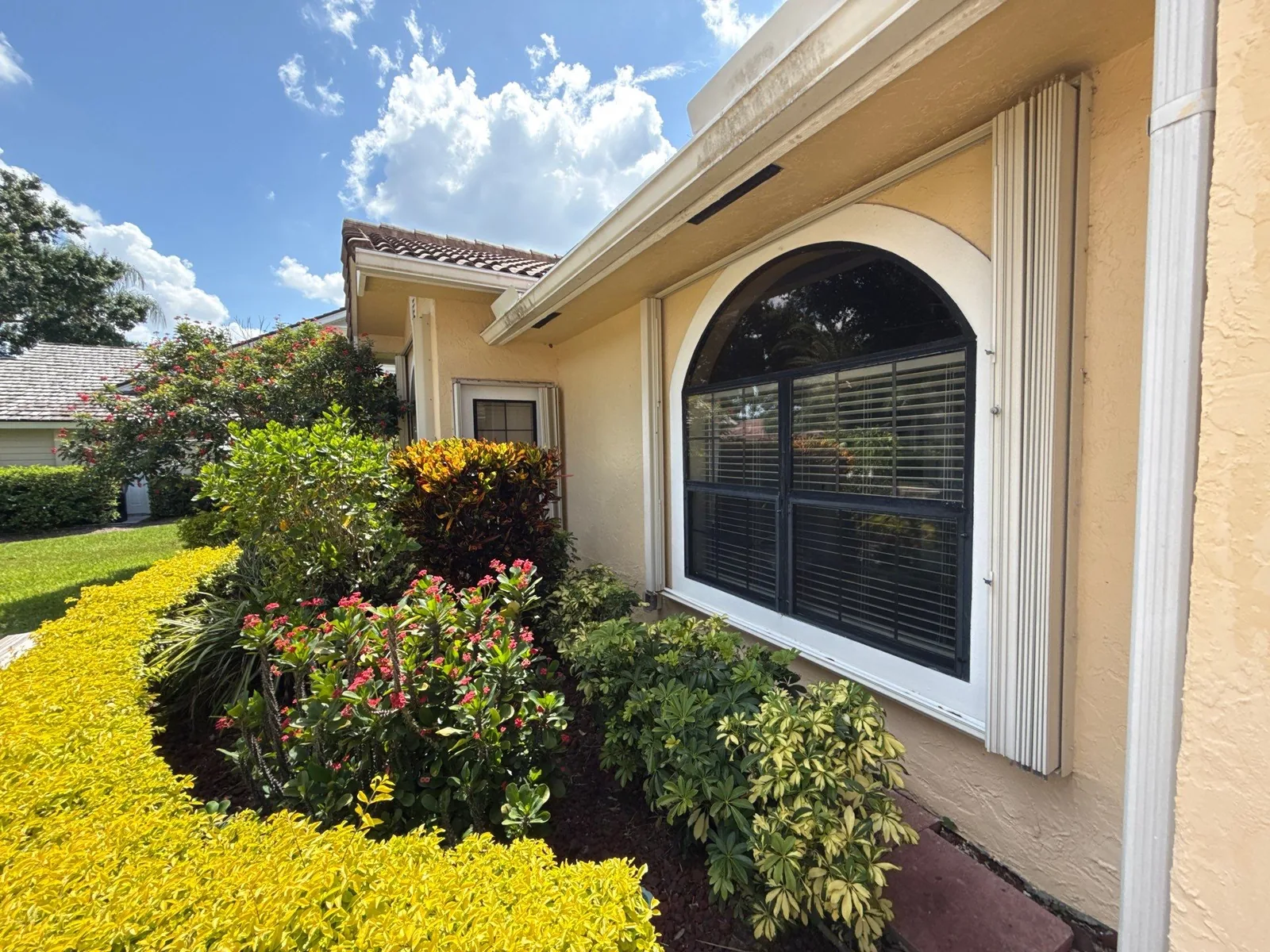 Exterior Painting in Port Saint Lucie, Florida. Yellow stucco single-storey home exterior with a white-trimmed arched window, tile roof and flowering landscaped bed under a blue sky.