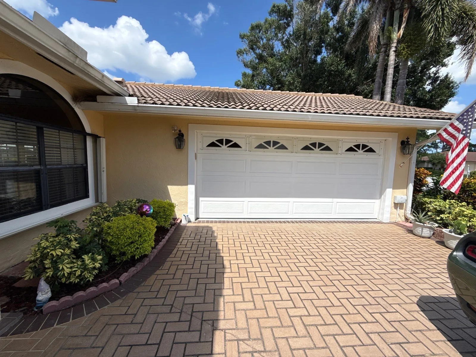 Exterior Painting in Port Saint Lucie, Florida. Front elevation of a yellow stucco home with a white two-car garage door, tile roof, paver driveway and landscaped border.
