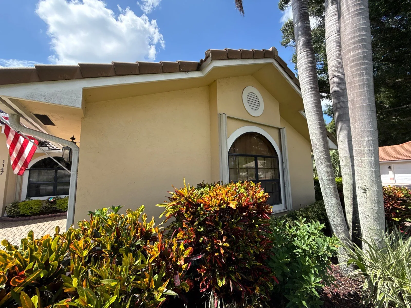 Exterior Painting in Port Saint Lucie, Florida. Yellow stucco gable end with a round vent, white-trimmed arched window and tile roof, framed by palms and croton shrubs.