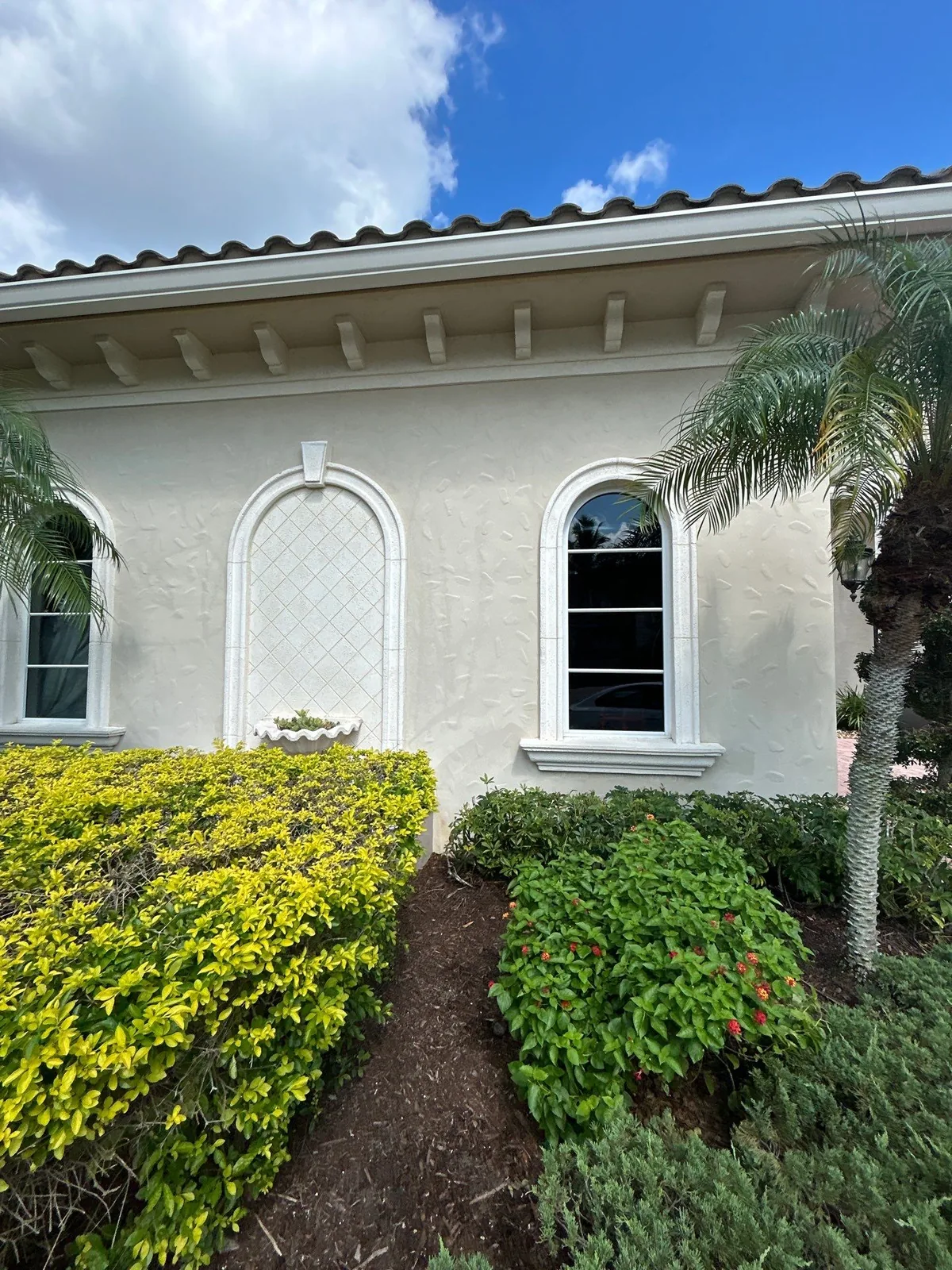 Exterior Painting in Port Saint Lucie, Florida. Single-storey stucco exterior wall painted off-white with white arched window trim, barrel tile roof and trimmed hedges under blue sky.