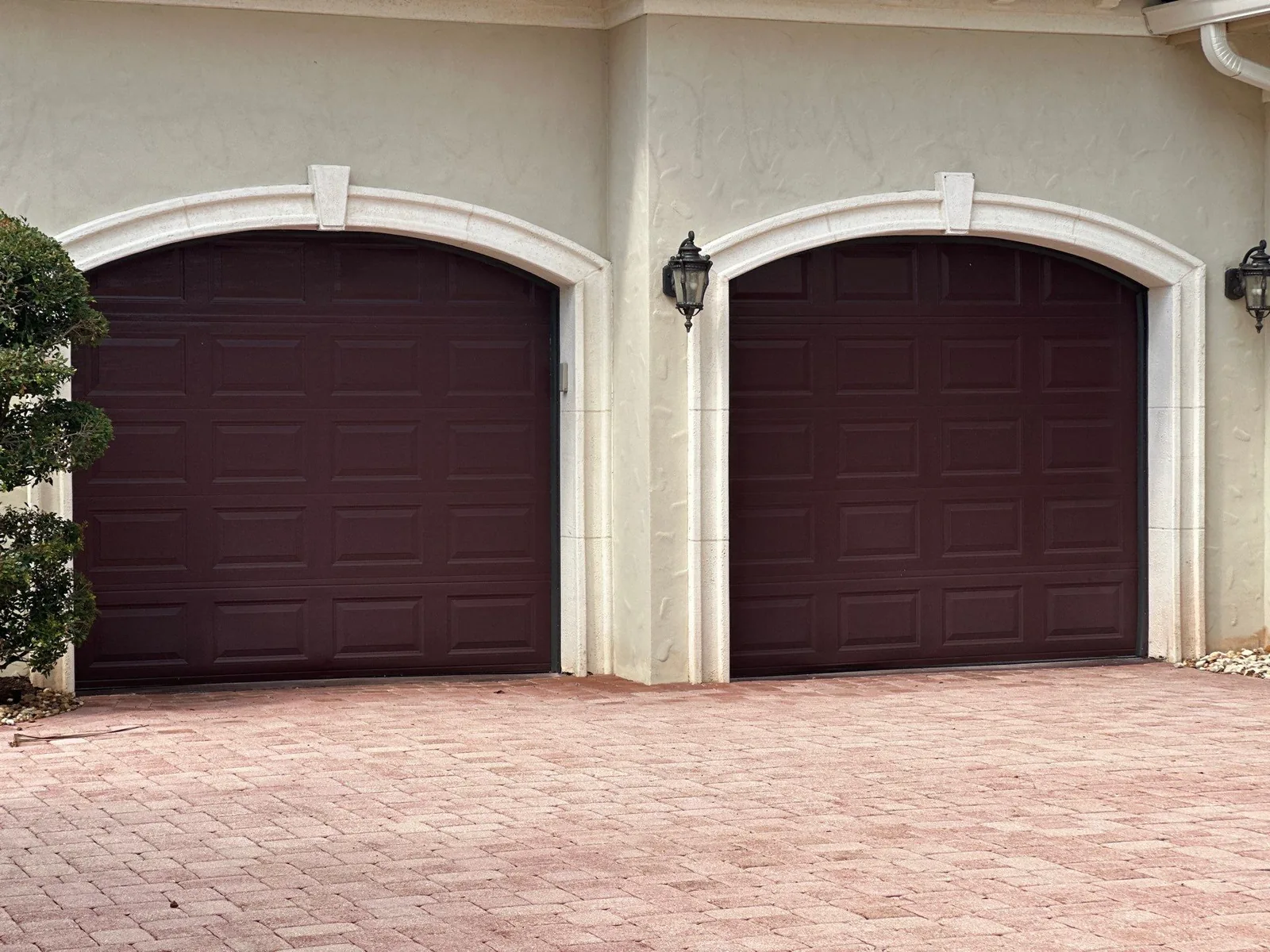 Exterior Painting in Port Saint Lucie, Florida. Two arched dark brown garage doors in a cream stucco wall with stone surrounds and coach lights, brick paver driveway.