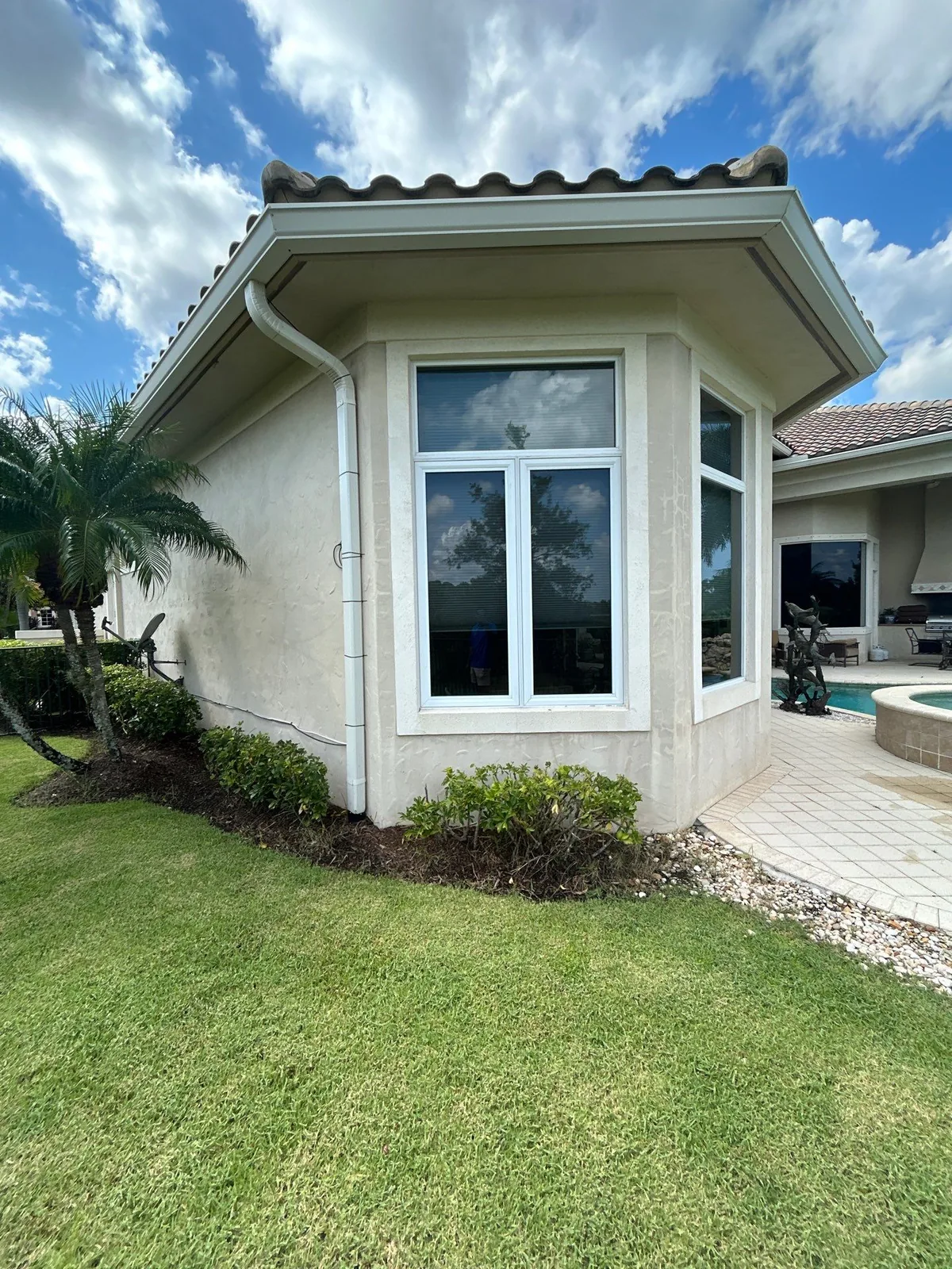 Exterior Painting in Port Saint Lucie, Florida. Corner of a single-storey wing with a bay window in white trim, cream stucco walls, tile roof, lawn and planting bed.