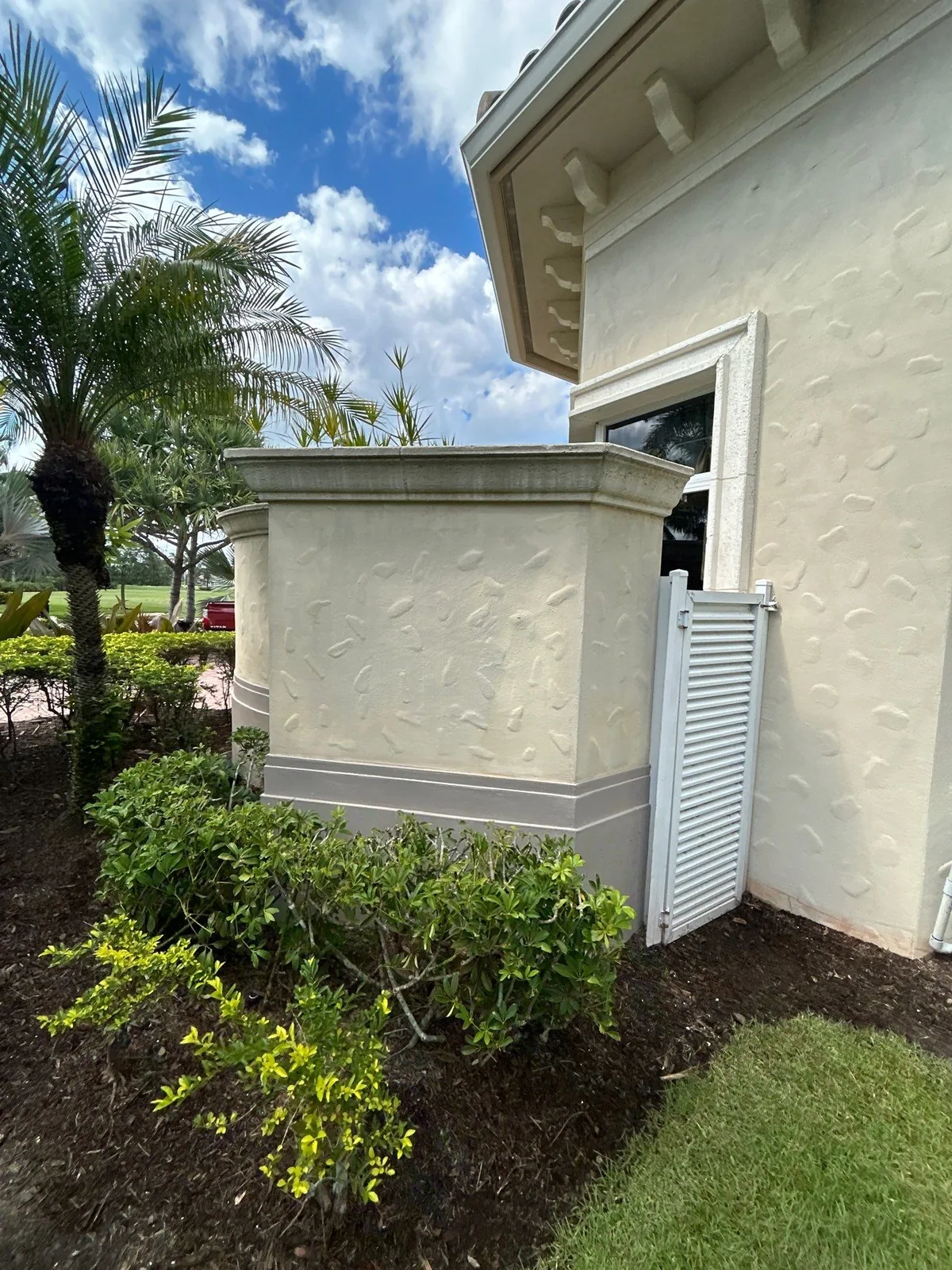 Exterior Painting in Port Saint Lucie, Florida. Stucco home exterior in cream with a grey base band and white trim, landscaped shrubs and palms under a blue sky.