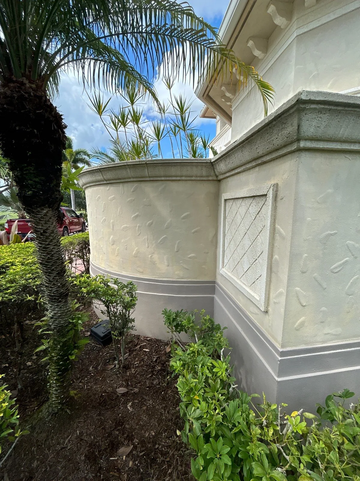 Exterior Painting in Port Saint Lucie, Florida. Curved stucco exterior wall in cream with a grey base band and a decorative lattice vent panel, palms and shrubs in front.