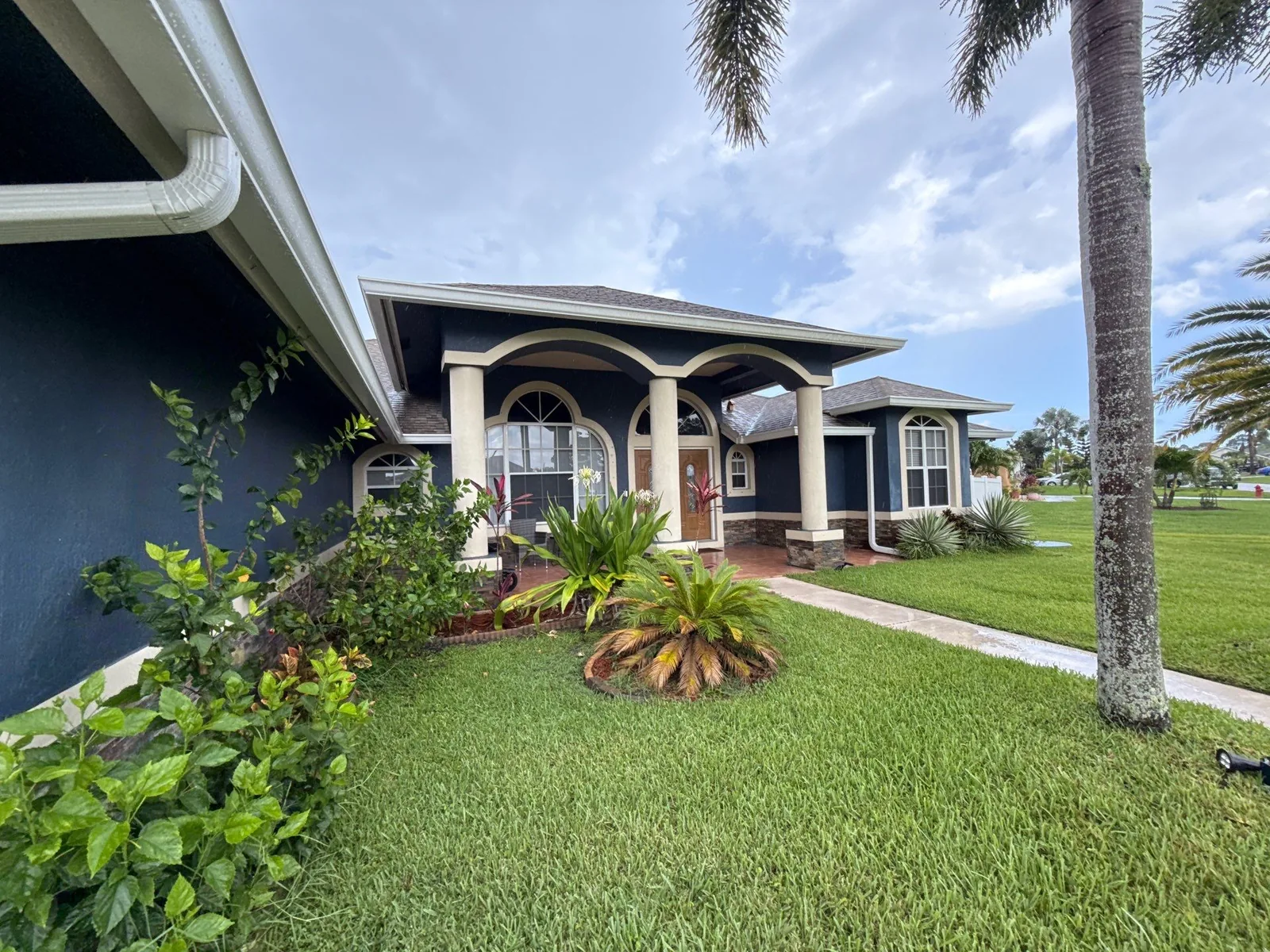 Exterior Painting in Port Saint Lucie, Florida. Front of a single-storey home with navy blue stucco walls and white trim, arched entry columns, tile roof and landscaped lawn.