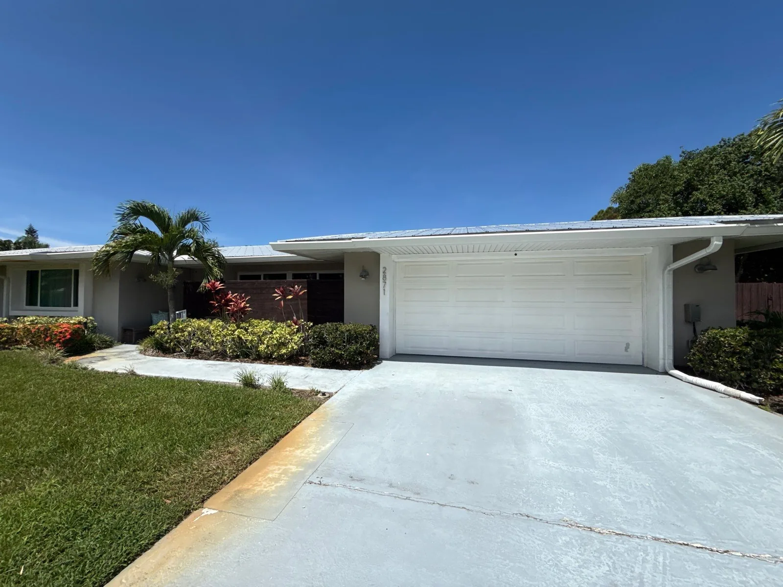Exterior Painting in Port Saint Lucie, Florida. Single-storey home painted white with a white garage door, a recessed entry with clerestory windows, clipped hedge, palm and concrete driveway.