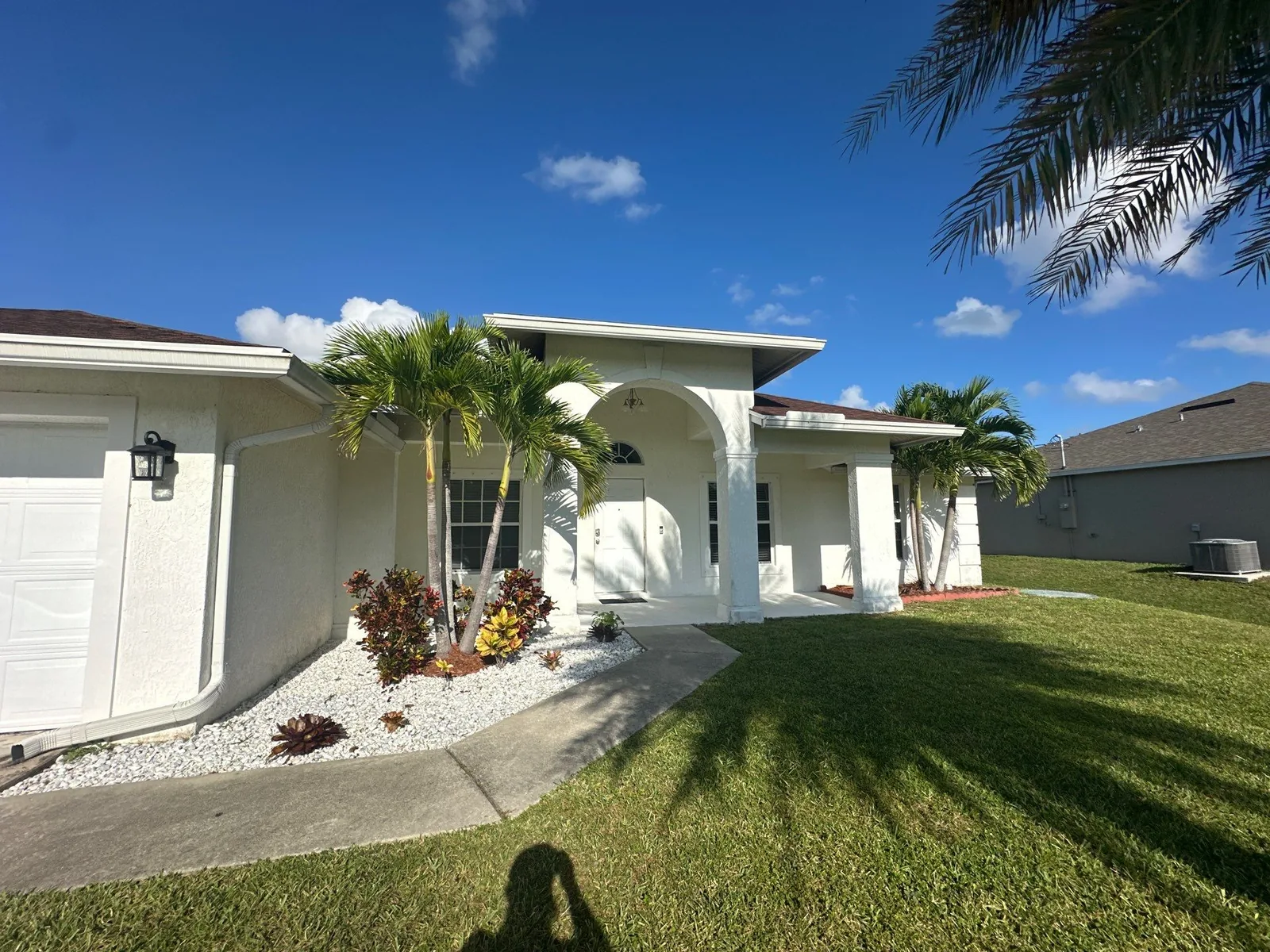 Exterior Painting in Port Saint Lucie, Florida. White stucco single-storey home with arched entry, palms and a stone planting bed under a clear blue sky.
