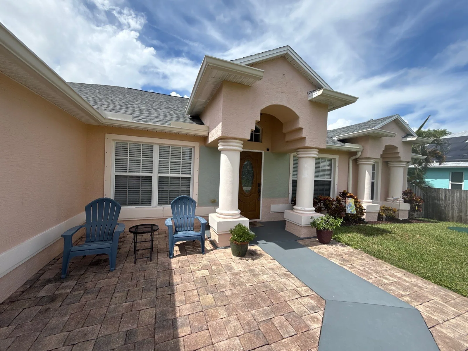 Exterior Painting in Port Saint Lucie, Florida. Front elevation of a single-storey home, pale pink stucco with white columns and arched entry, wood door and paver driveway.