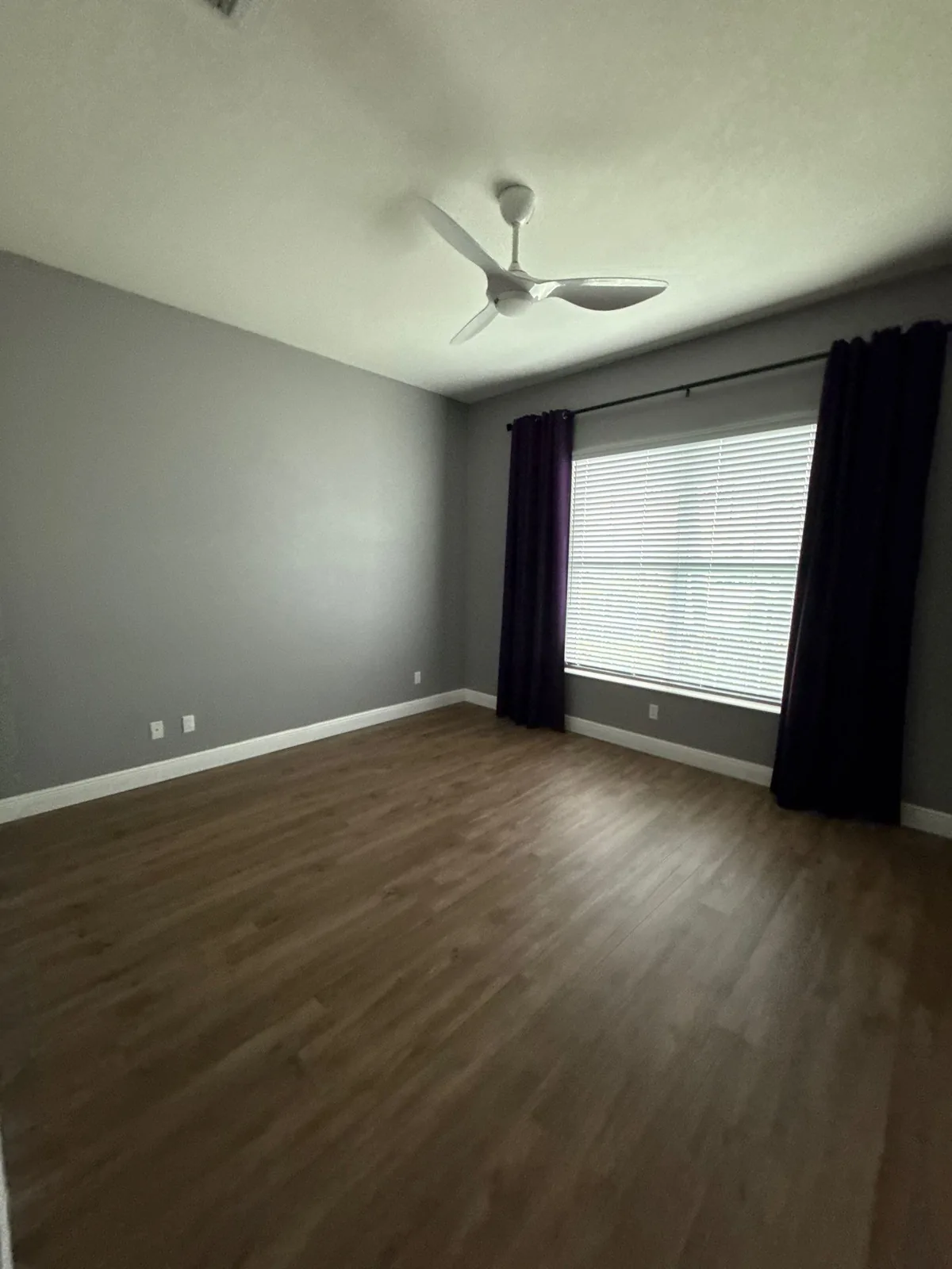 Interior Painting in Port Saint Lucie, Florida. Empty bedroom with grey painted walls, white ceiling and baseboards, dark curtains and wood-look flooring.