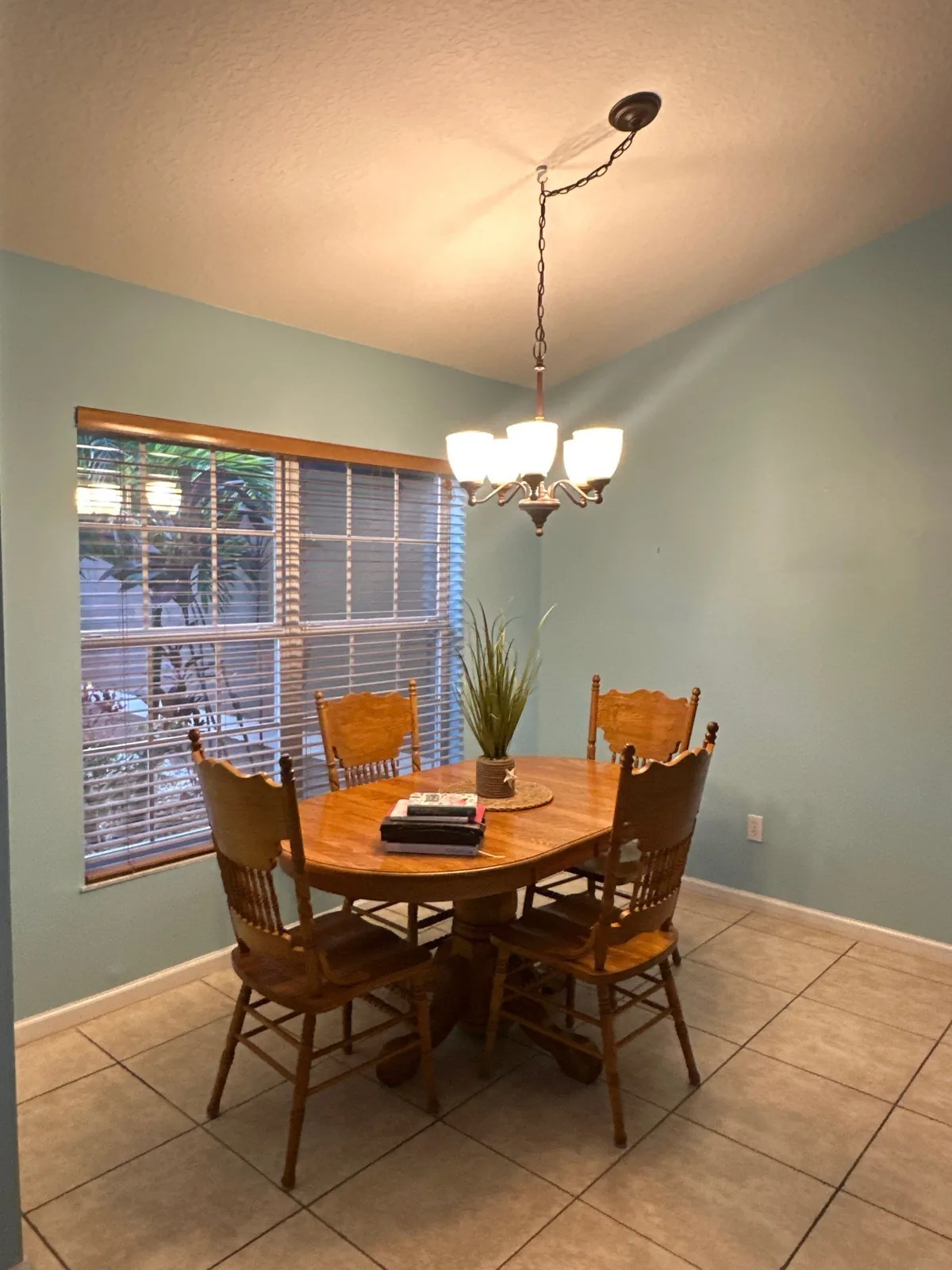 Interior Painting in Port Saint Lucie, Florida. Dining area with pale green walls, wood table and four wood chairs under a chandelier, blinded window.