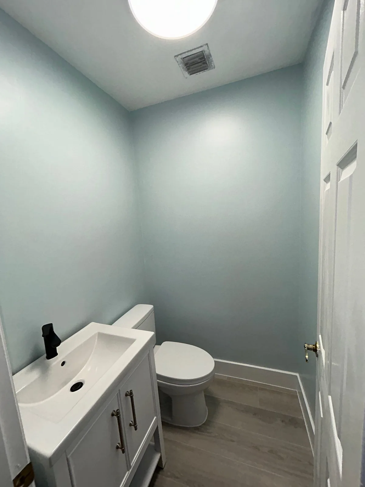 Interior Painting in Port Saint Lucie, Florida. Small powder room with pale blue painted walls and white ceiling, white vanity with black faucet and a toilet.
