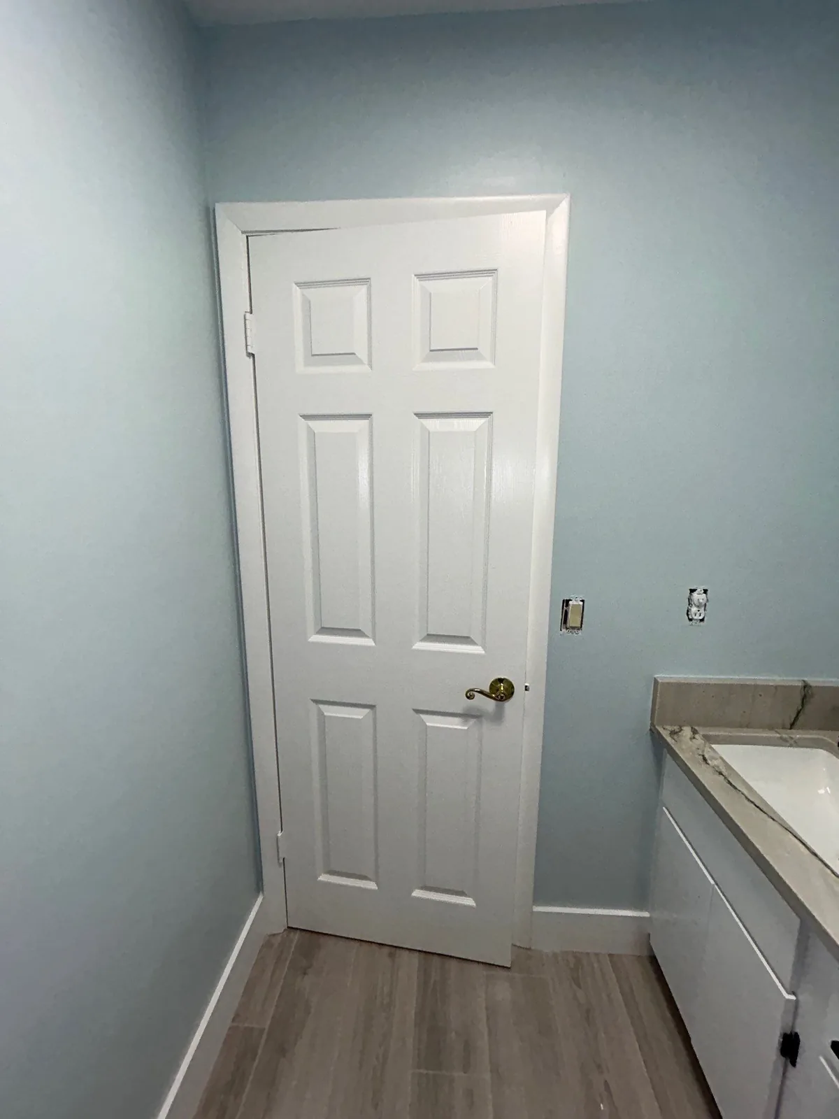 Interior Painting in Port Saint Lucie, Florida. Pale blue bathroom wall with a freshly painted white six-panel door, white baseboard and edge of a vanity.