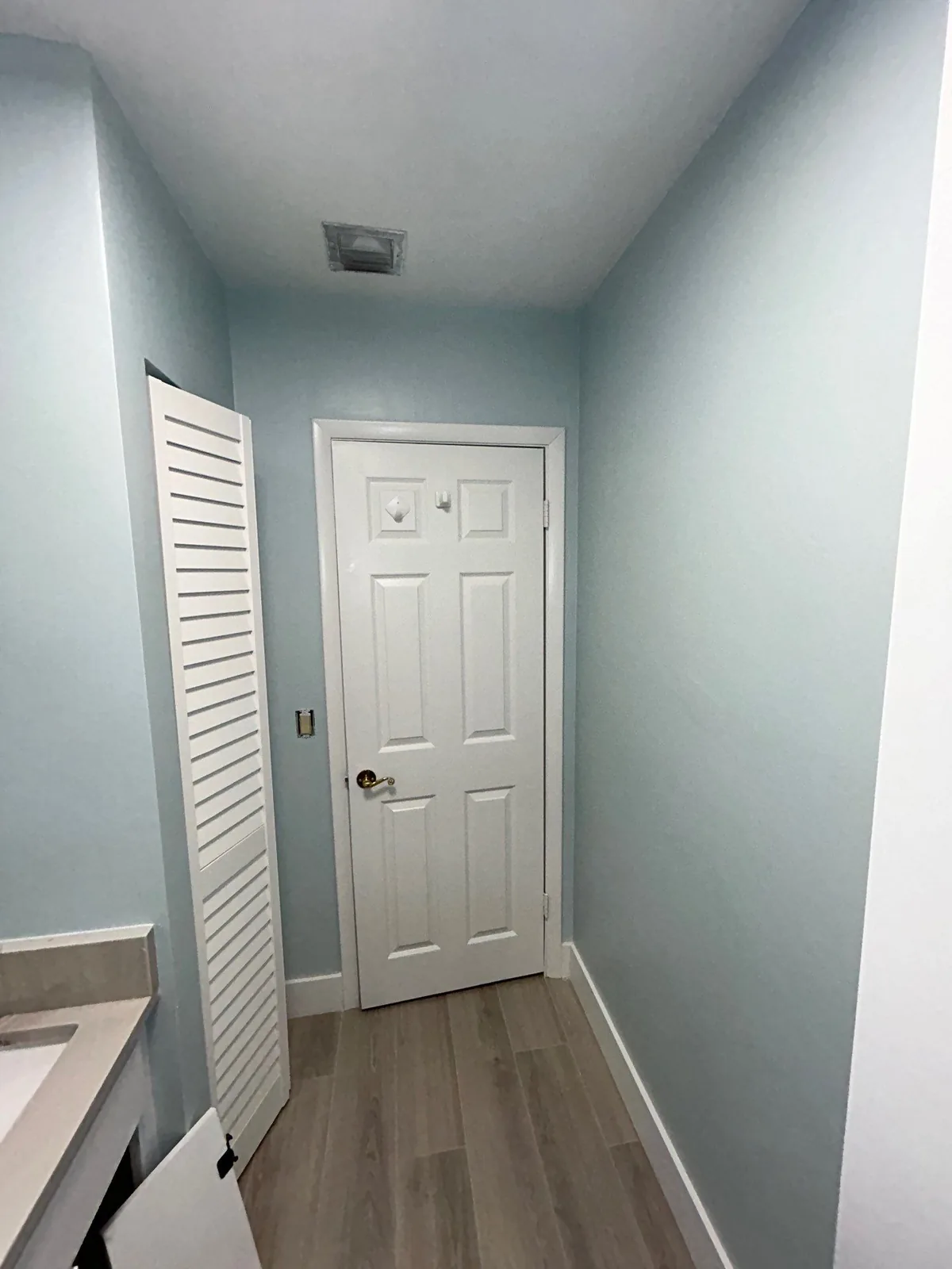 Interior Painting in Port Saint Lucie, Florida. Narrow bathroom passage with pale blue walls, a white six-panel door and a white louvred door, grey plank flooring.