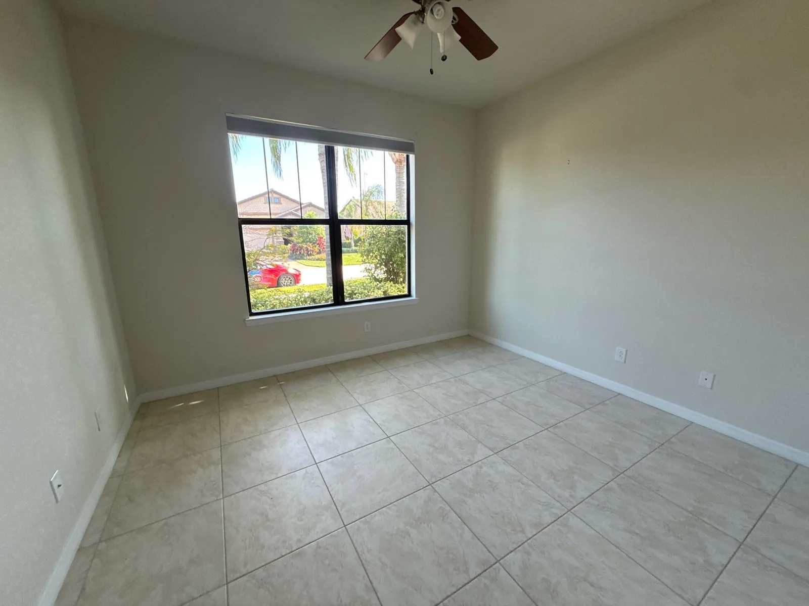 Interior Painting in Port Saint Lucie, Florida. Empty bedroom with light grey painted walls, tile floor, ceiling fan and a window onto greenery.