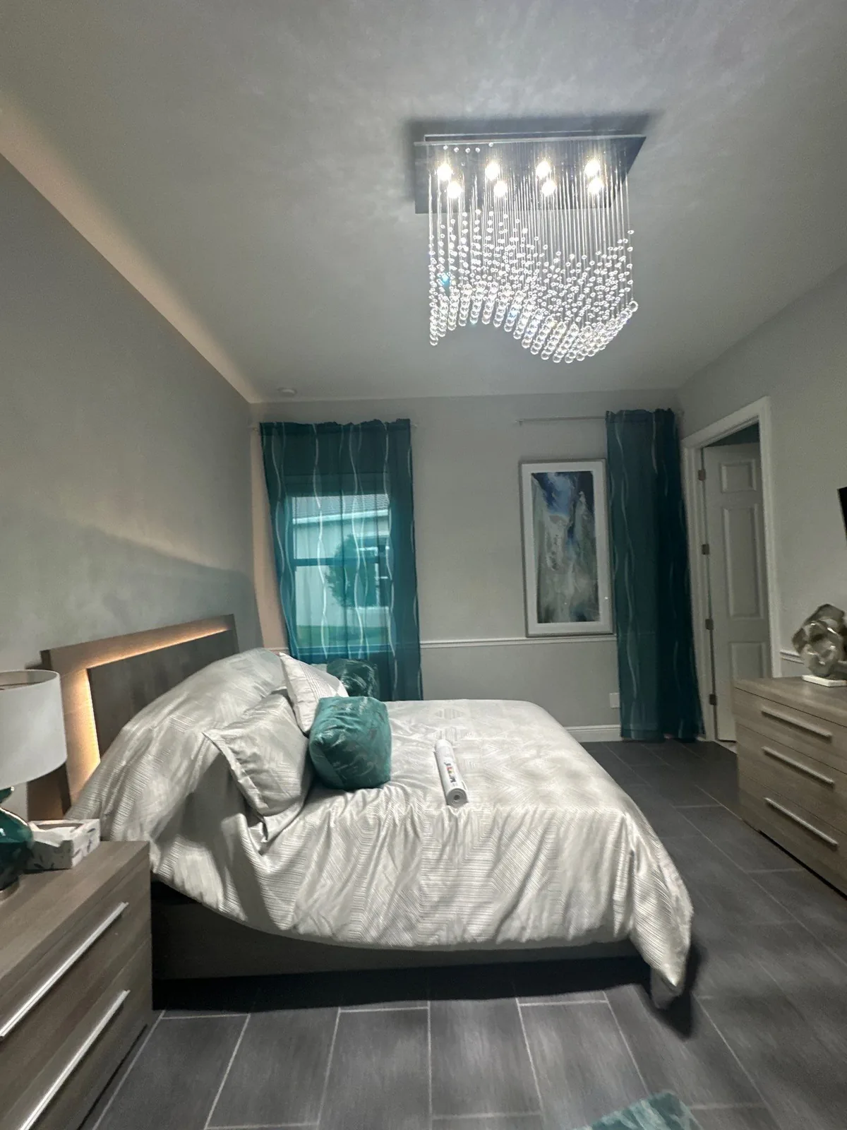 Interior Painting in Port Saint Lucie, Florida. Furnished bedroom with grey painted walls and white ceiling, teal curtains, crystal chandelier and grey tile floor.