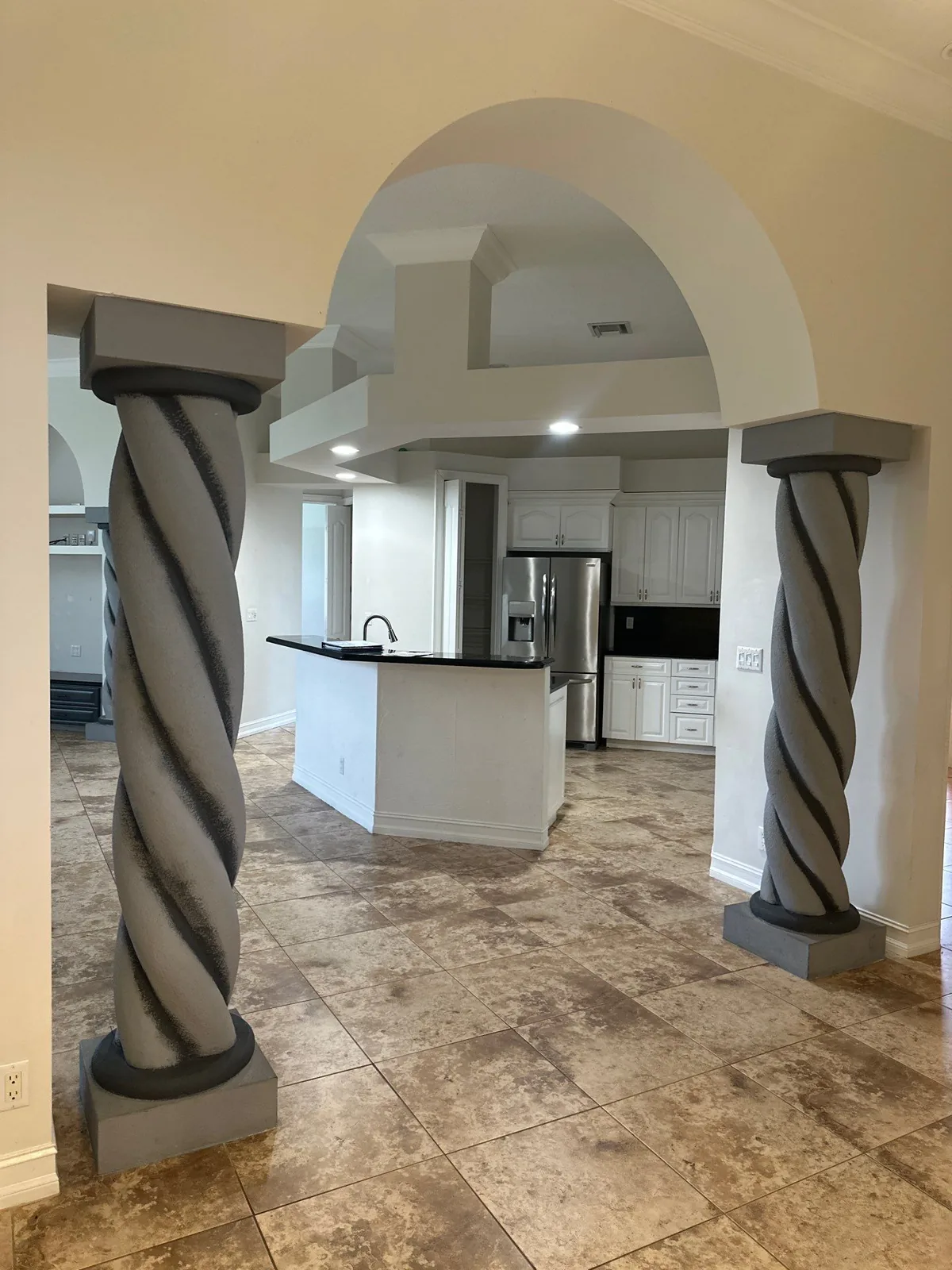 Interior Painting in Port Saint Lucie, Florida. Interior archway framed by two spiral-carved decorative columns painted grey, with a kitchen with white cabinets, dark countertop island and stainless refrigerator beyond.