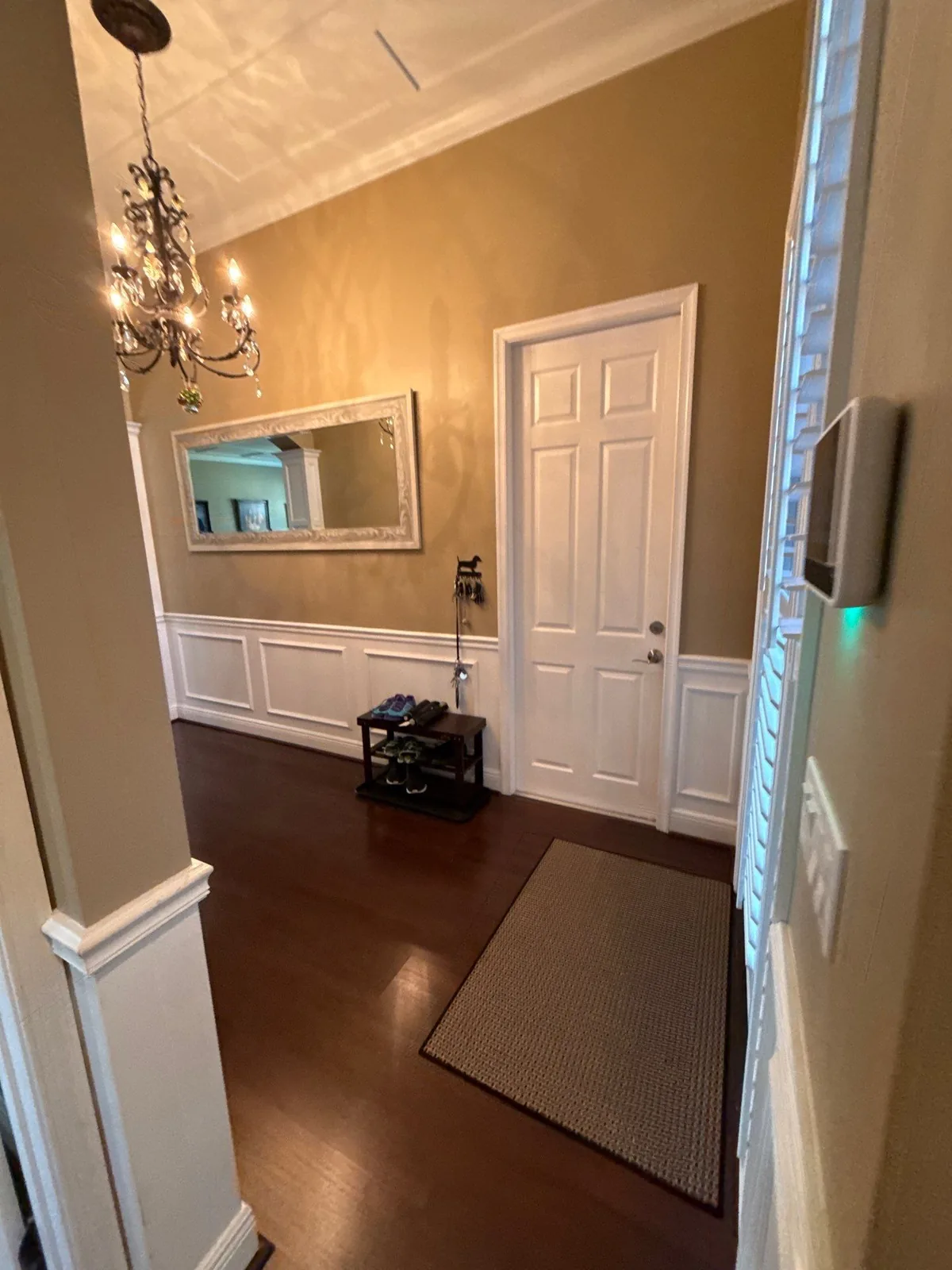 Interior Painting in Port Saint Lucie, Florida. Entry hallway with tan walls above white raised-panel wainscoting, white crown moulding and six-panel door, chandelier and framed mirror over dark wood flooring.