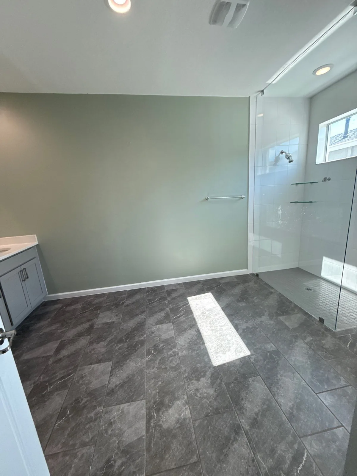 Interior Painting in Port Saint Lucie, Florida. Bathroom with a sage green painted wall, glass walk-in shower with white tile and grey vanity over dark tile flooring.