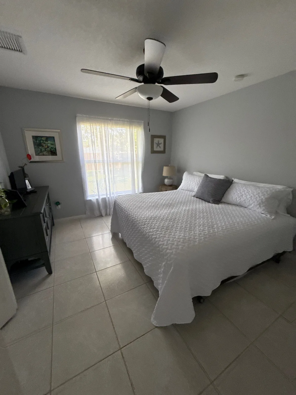 Interior Painting in Port Saint Lucie, Florida. Bedroom with light grey walls and white ceiling, dark ceiling fan, a made king bed with white quilt and grey pillows, and a dark dresser.