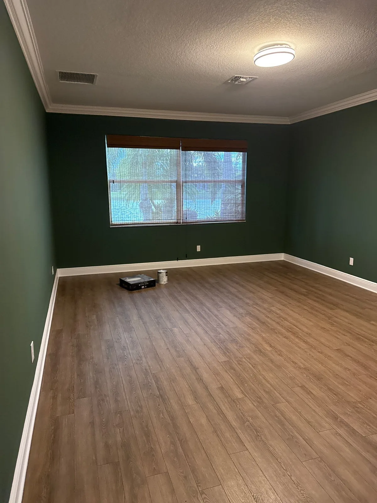 Interior Painting in Port Saint Lucie, Florida. Empty bedroom with dark green walls, white baseboards and crown moulding, wood-look plank flooring and a window.