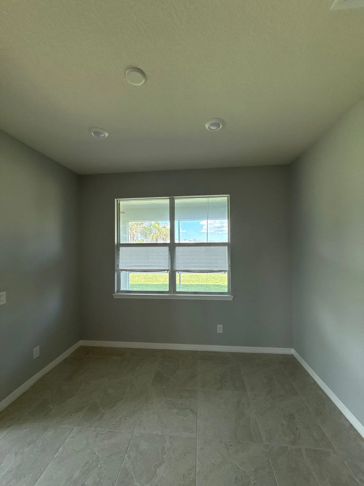 Interior Painting in Port Saint Lucie, Florida. Empty room with gray walls, white baseboards and a white-trimmed window with blinds over tile flooring.