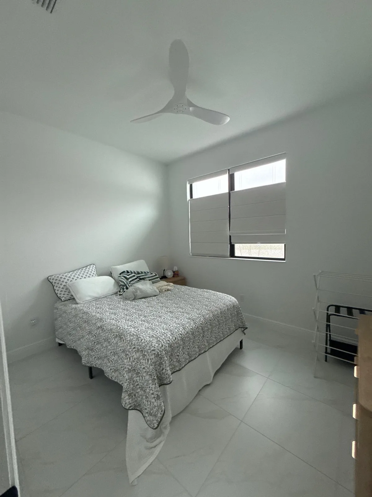 Interior Painting in Port Saint Lucie, Florida. Bedroom with white walls and ceiling, ceiling fan, made bed and window with blinds over tile floor.