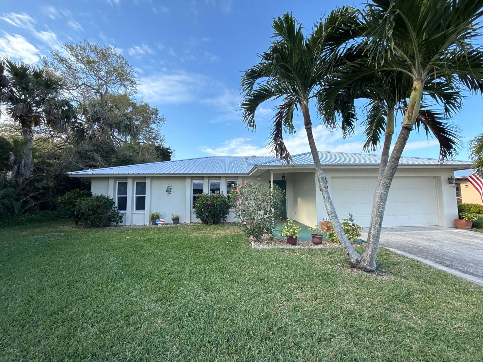 Exterior Painting in Port Salerno, Florida. Front elevation of a single-storey beige house with white garage door, metal roof, palm trees and mown lawn.
