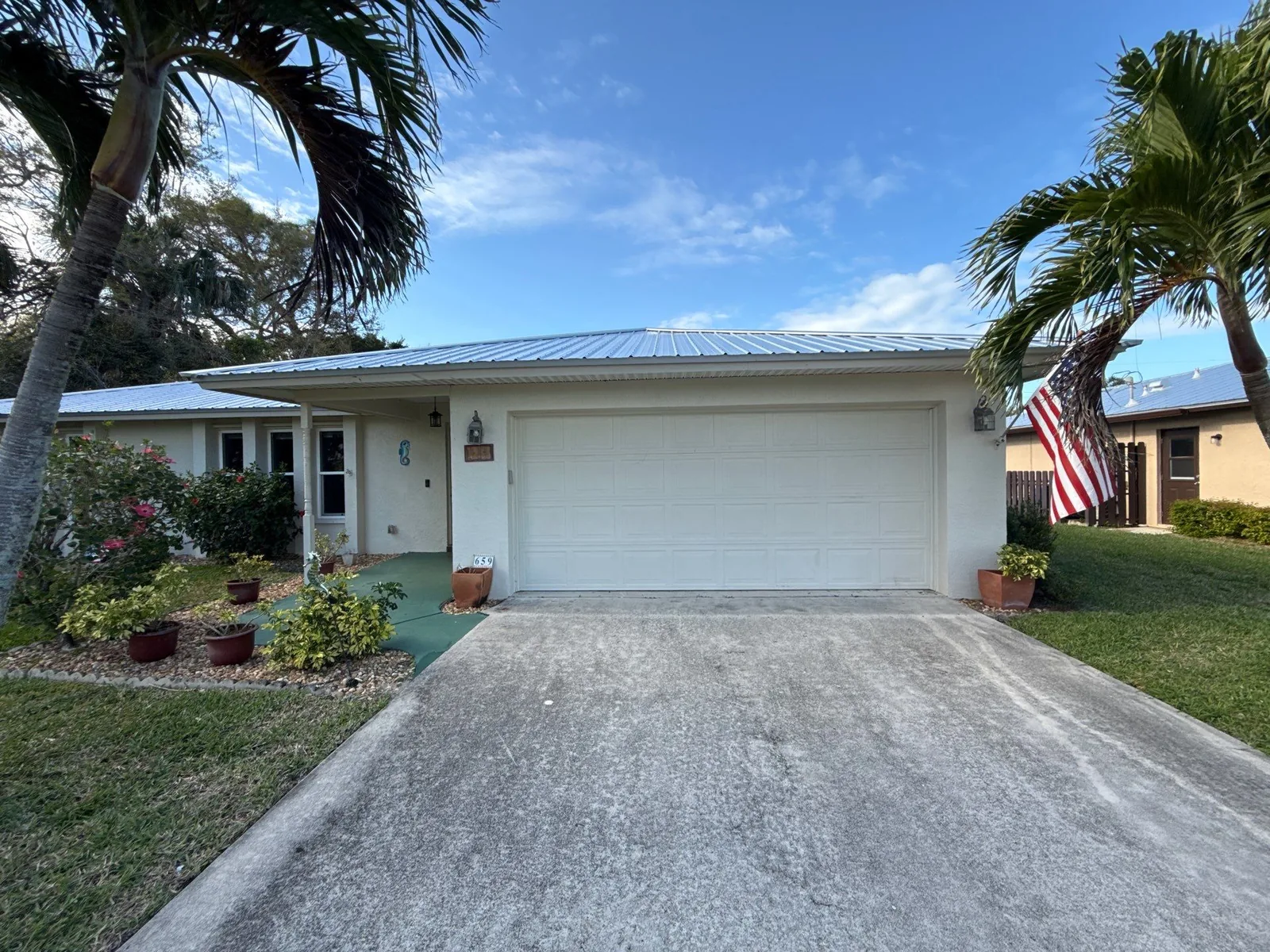 Exterior Painting in Port Salerno, Florida. Front elevation of a single-storey white house with a metal roof, white garage door and palm trees, in daylight.