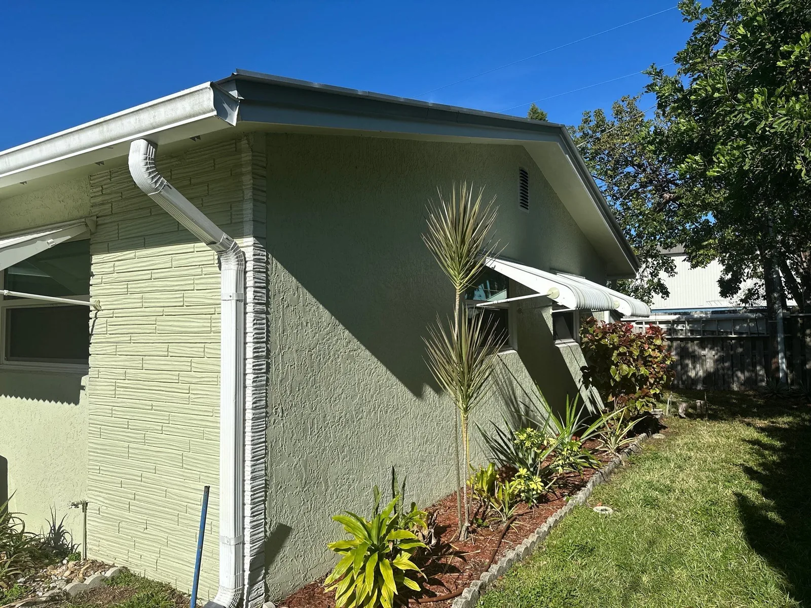 Exterior Painting in Port Salerno, Florida. Corner of a pale green stucco house with white fascia, gutter and downspout, planting bed alongside.
