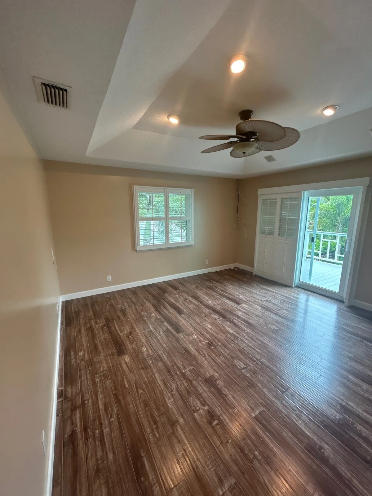 Interior Painting in Port Salerno, Florida. Empty bedroom with beige walls, white trim, tray ceiling, ceiling fan and plantation-shutter doors to a deck.