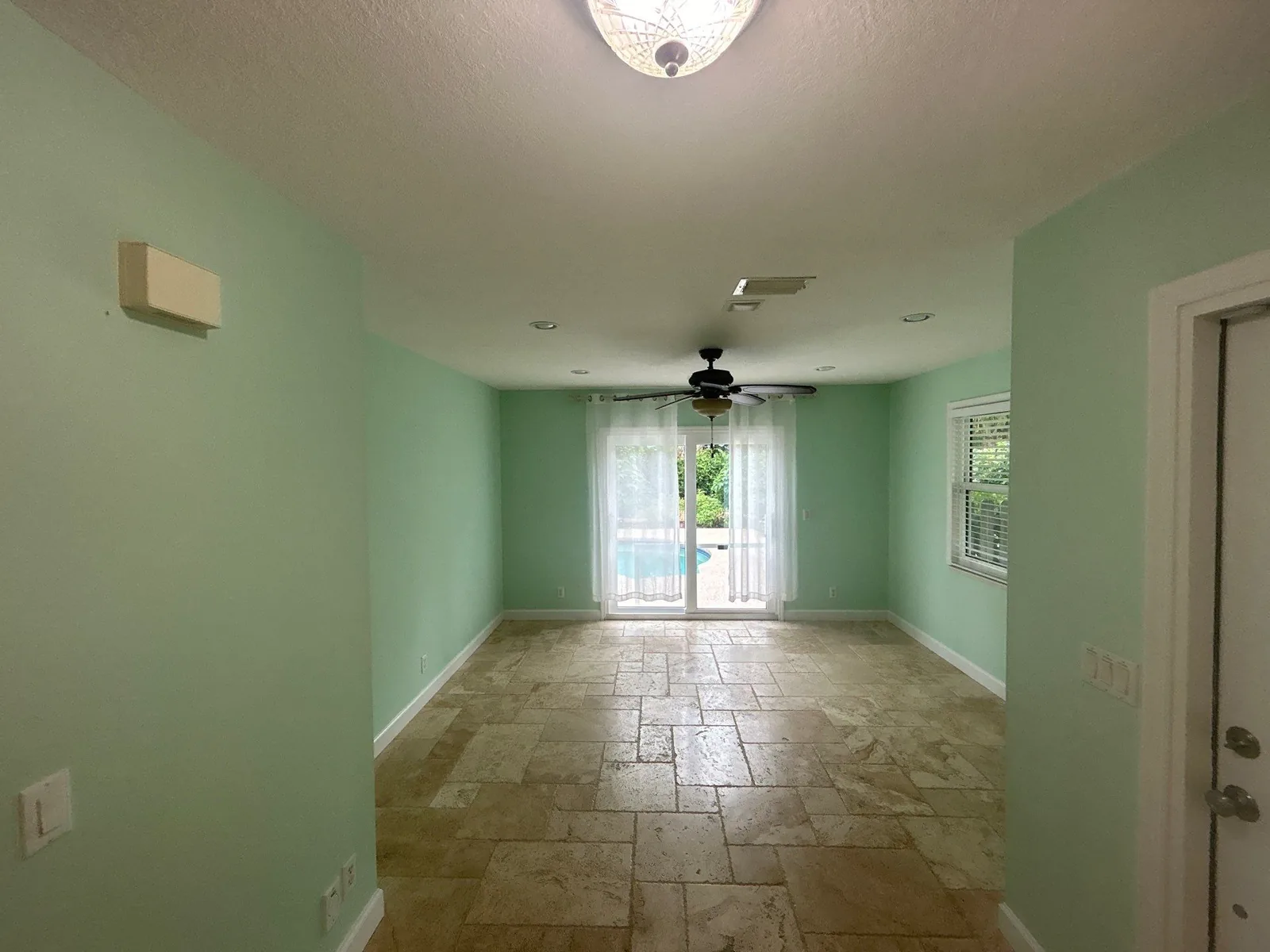 Interior Painting in Port Salerno, Florida. Empty living room with mint green walls, white trim and baseboards, stone tile floor and sliding glass doors to a pool.