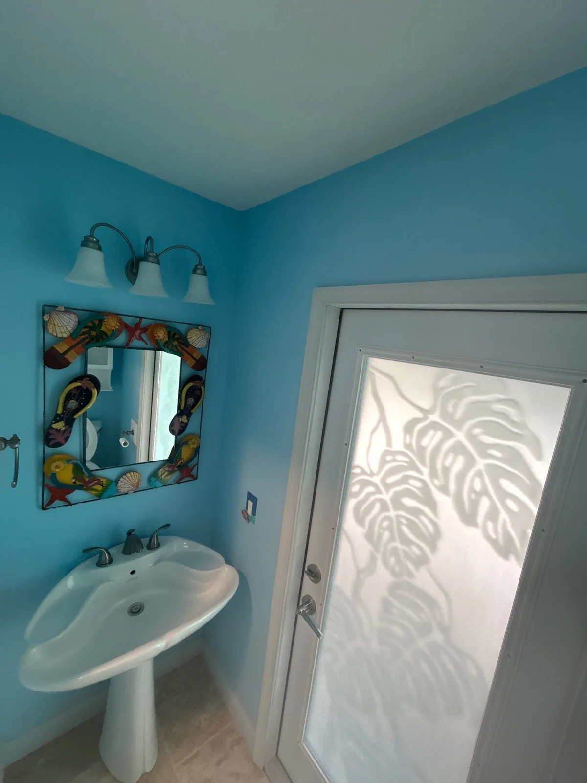 Interior Painting in Port Salerno, Florida. Half bathroom with blue walls, white pedestal sink, three-light vanity fixture, decorative mirror and a frosted glass door with a leaf pattern.