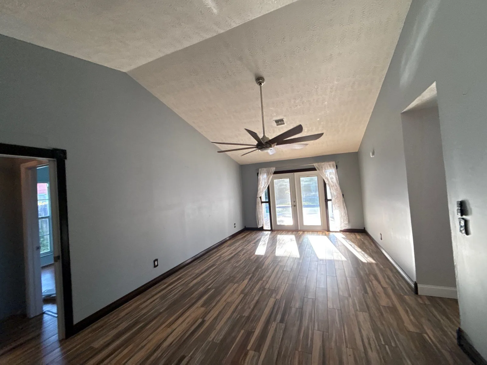 Interior Painting in Port Salerno, Florida. Empty living room with grey painted walls, white textured vaulted ceiling, ceiling fan and french doors with daylight across wood-look flooring.