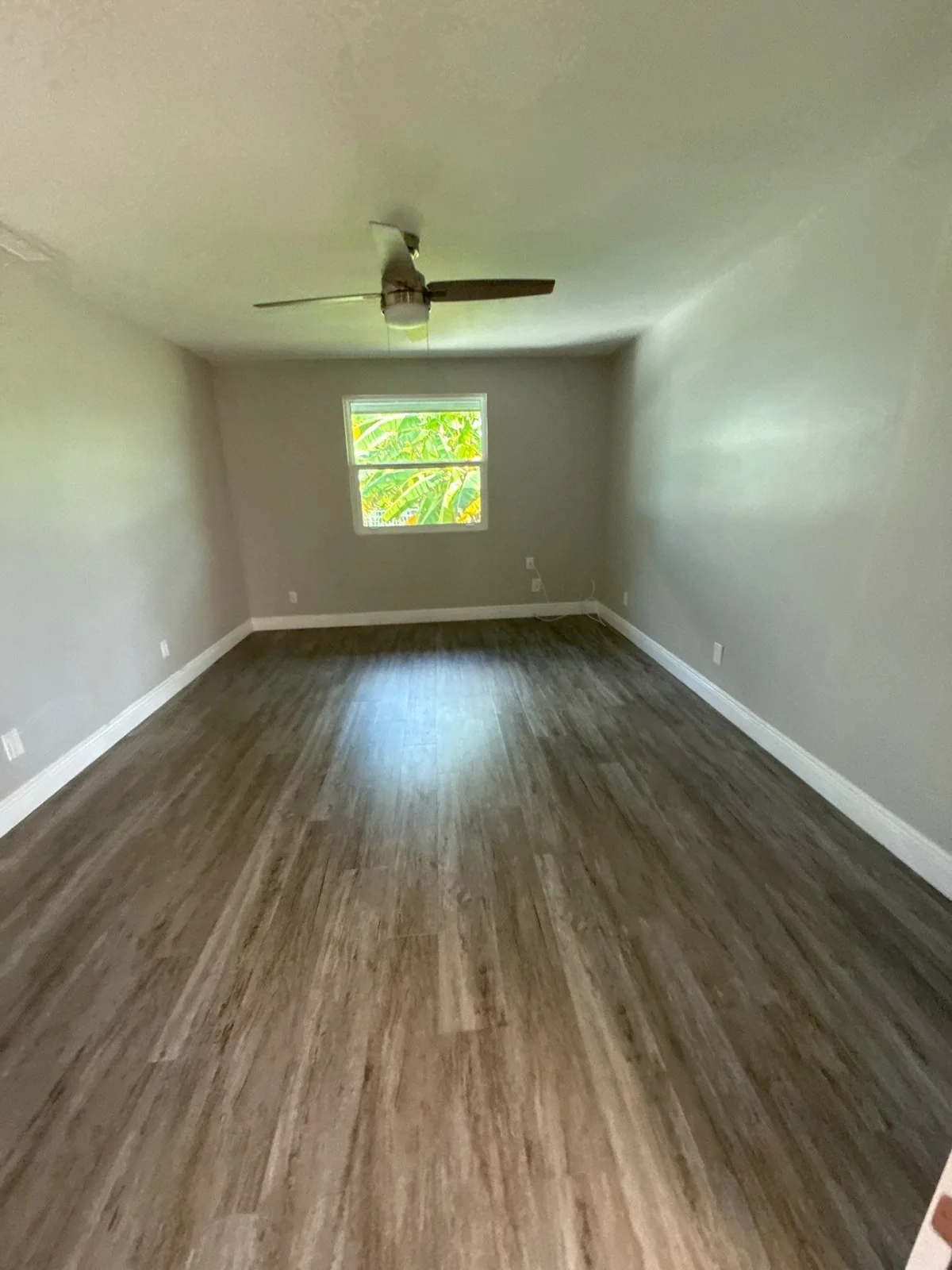 Interior Painting in Port Salerno, Florida. Empty bedroom with grey walls, white ceiling and baseboards, ceiling fan, single window and dark wood-look plank flooring.