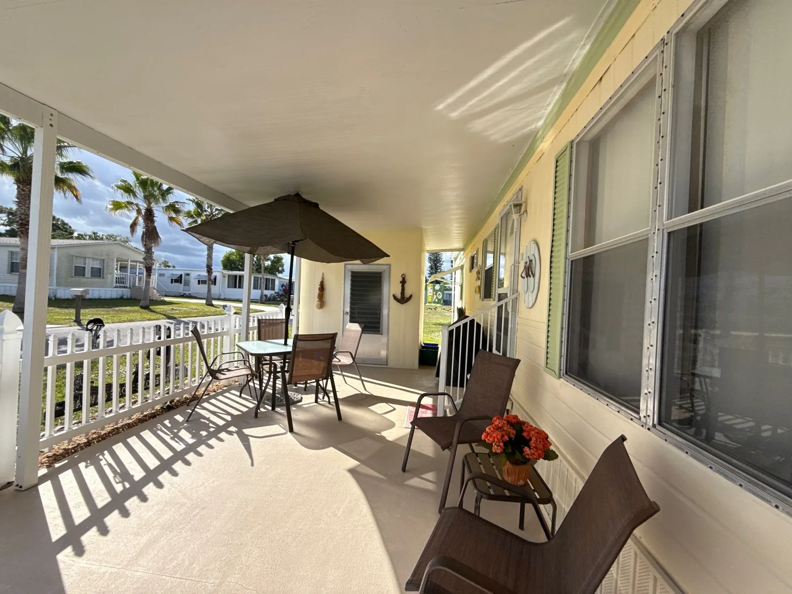 Exterior Painting in River Park, Florida. Covered porch of a manufactured home with pale yellow painted siding, white ceiling and railing, patio table and chairs, palms and blue sky beyond.